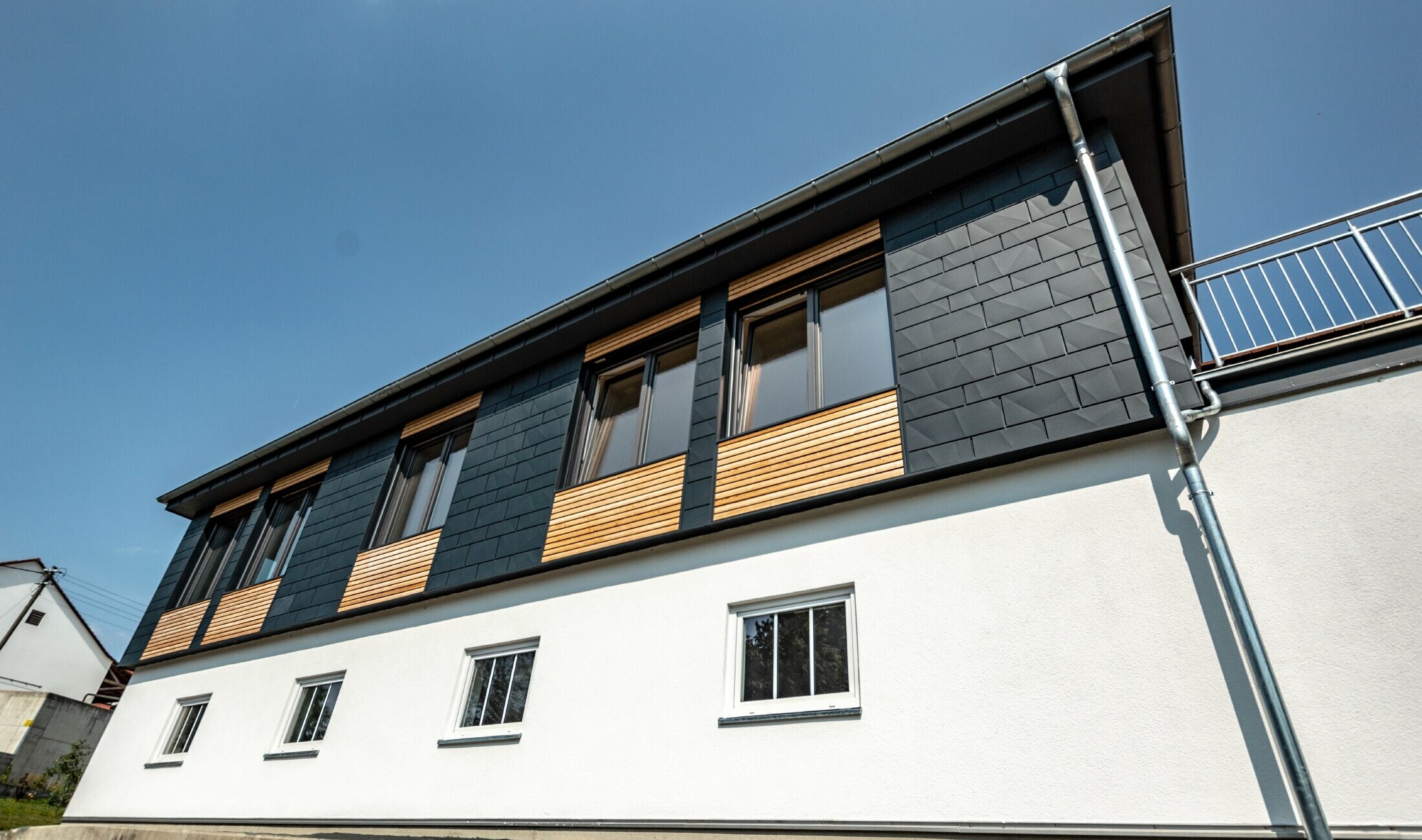 Conception de façade avec différents matériaux de construction : Siding.X PREFA en aluminium couleur anthracite, habillage en bois horizontal et façade en crépi blanc.