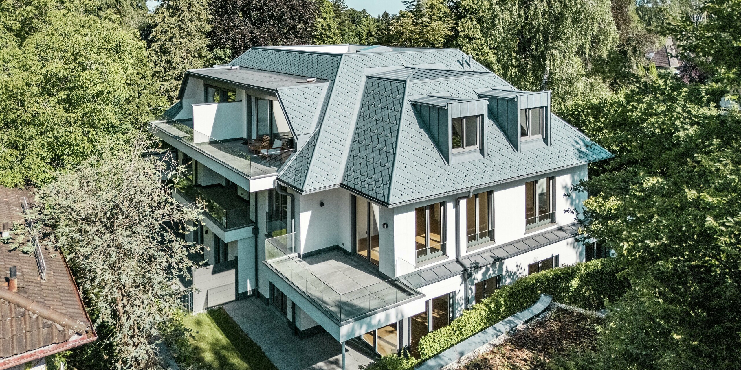 Vue aérienne d'un immeuble d'appartements moderne à la toiture au design remarquable, recouverte de losanges PREFA 29 x 29 cm dans l'élégant coloris Gris Pierre P.10. L'imposante toiture mansardée, associée aux élégantes lucarnes PREFALZ, crée un effet visuel saisissant. Les produits en aluminium de haute qualité s'intègrent harmonieusement au paysage verdoyant et offrent des avantages à la fois esthétiques et fonctionnels. La précision de fabrication et la résistance aux intempéries des produits PREFA garantissent leur durabilité et protègent le bâtiment des éléments.