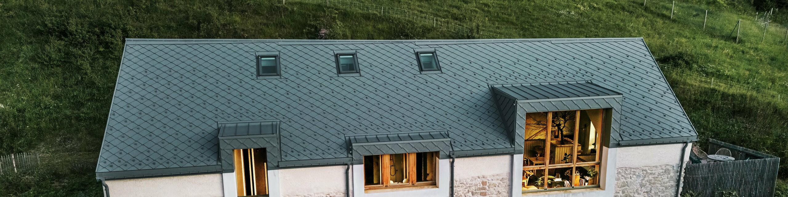 Vue nocturne de la ferme restaurée "La Grange à Gaby" à Engins, France. Le bâtiment combine la maçonnerie en pierre traditionnelle avec un toit moderne en aluminium PREFA composé de losanges de toiture de 29 × 29 cm. Les grandes fenêtres et les intérieurs illuminés soulignent l’atmosphère accueillante du nouveau centre culturel.