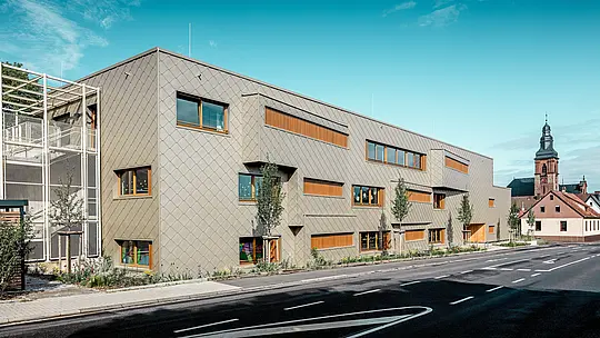 Baignée de lumière naturelle, la crèche moderne d'Alzenau dévoile avec éclat sa remarquable façade en aluminium PREFA, composée de losanges de façade 44 x 44 couleur bronze. La structure uniforme, semblable à des écailles, ponctuée de fenêtres décalées et de persiennes verticales en bois, confère à la façade profondeur et rythme. À gauche, on aperçoit la cage d'escalier délicate, avec sa structure métallique en grille, qui contribue à l'impression de transparence et de légèreté. À l'arrière-plan se dresse l'église gothique tardive d'Alzenau, symbole de la fusion harmonieuse entre traditions architecturales historiques et architecture contemporaine. Le système de façade PREFA, durable et sans entretien, soutient la conception novatrice, fonctionnelle et esthétiquement aboutie de ce bâtiment éducatif tourné vers l'avenir.