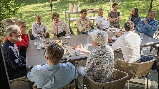 PREFARENZEN 2027 : Dialogue d’experts dans le jardin luxuriant d’un domaine viticole : architectes et designers, réunis autour d’une grande table, échangent des points de vue passionnants sur l’architecture moderne. La lumière du soleil, filtrée par les arbres, teinte les visages et les vêtements d’une douce clarté. Verres, bouteilles d’eau et en-cas ornent la table, tandis qu’à l’arrière-plan, les produits en aluminium PREFA, aux lignes épurées et à la robustesse reconnue, sont mis en valeur. Cette scène illustre comment les toitures et façades PREFA conjuguent design, fonctionnalité et esthétique durable, et stimule la créativité.