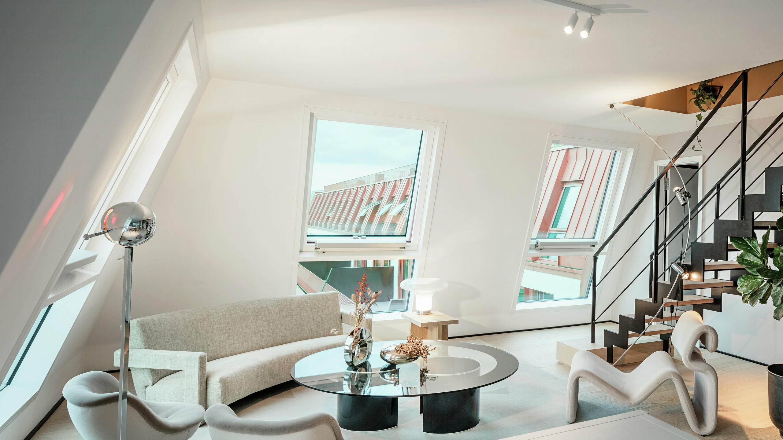 Vue intérieure d'un penthouse baigné de lumière dans le projet résidentiel Van B à Munich avec vue sur le toit en aluminium PREFALZ de haute qualité dans la couleur spéciale IGP Orange HDP. Les grandes fenêtres laissent entrer beaucoup de lumière du jour et soulignent le concept de vie moderne. Des lignes claires, des meubles design et une finition précise des raccords de toit montrent comment une architecture de haute qualité et élégante peut être mise en œuvre non seulement à l'extérieur mais aussi à l'intérieur.