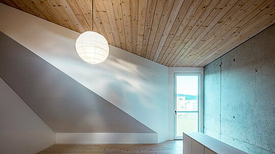L'image présente une pièce minimaliste dans une maison individuelle à Freistadt. Le plafond en sapin blanc confère à la pièce une atmosphère chaleureuse et accueillante, accentuée par la simplicité du design et la lumière naturelle qui pénètre par la fenêtre de droite. Cette fenêtre offre une vue sur la verdure environnante et renforce la sensation d'espace. La pièce se caractérise par des murs lisses et neutres. L'alliance du mur en béton et du plafond en bois crée un contraste harmonieux, empreint d'élégance et de naturel. L'aménagement intérieur soigné reflète la philosophie de toute la maison : fonctionnalité, clarté esthétique et matériaux intemporels.