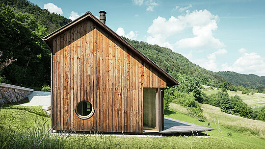 Maison en bois minimaliste avec une ouverture de fenêtre ronde et un toit en aluminium PREFALZ durable de couleur P.10 brun, nichée dans le paysage vallonné de Vinje. La couverture de toit PREFA, sans entretien, offre une protection optimale contre le vent et les intempéries, tout en s’accordant parfaitement avec la façade en bois naturel. Elle met en valeur l’architecture claire et intemporelle du bâtiment. Depuis la vaste terrasse, on profite d’une vue imprenable sur les collines slovènes. La perspective montre la façade latérale de cette maison individuelle avec son toit à deux pans asymétrique et ses surfaces de toit de tailles différentes.
