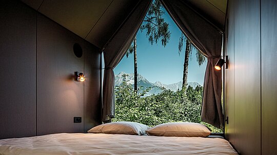 Vue depuis le lit d'une tiny house minimaliste dans le Secret Koi Garden, au bord du lac Natterer : la grande baie vitrée triangulaire ouvre la pièce sur la nature environnante, avec ses arbres et la vue sur les montagnes tyroliennes. L'intérieur épuré invite à la contemplation et souligne l'importance de ralentir le rythme. L'architecture est protégée par des losanges en aluminium PREFA, un matériau durable qui allie design, résistance aux intempéries et durabilité.