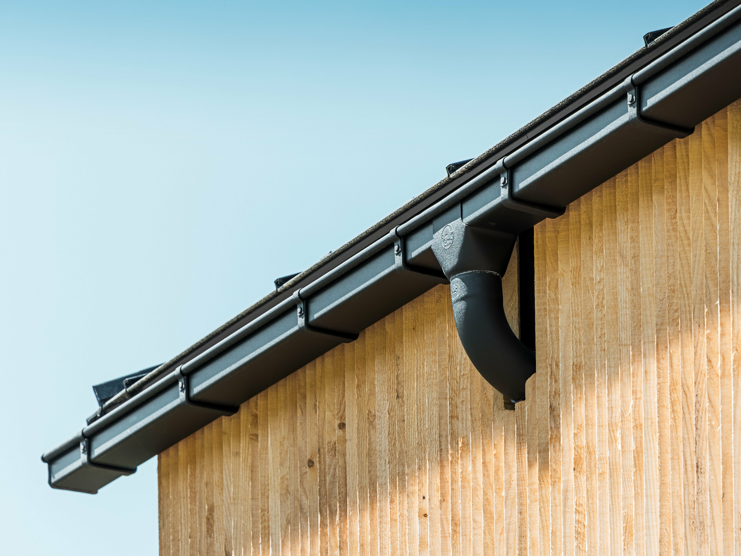 Angle de maison avec gouttière carrée PREFA en noir, avec caisson, fixation à l'aide de crochets de gouttière PREFA, façade revêtue de bois.