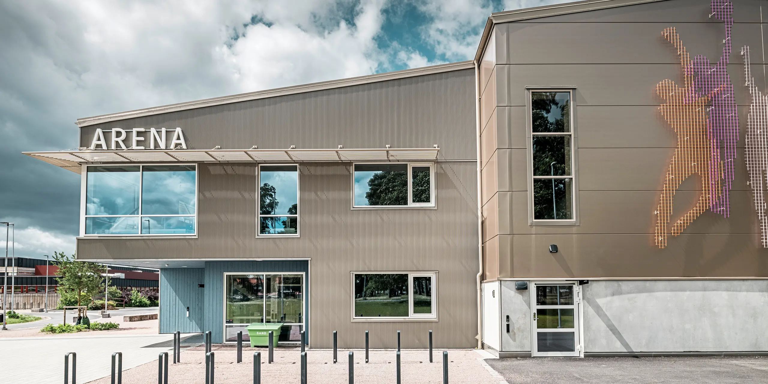 Vue latérale de la zone d'entrée du Park Arena de Värnamo, en Suède, avec une façade PREFA moderne constituée d'un profil triangle en bronze. La façade en aluminium est durable et crée des effets d’ombre et de lumière saisissants. À droite de l'image se trouve une fresque murale de conception artistique qui représente des personnages sportifs et souligne l'atmosphère dynamique de la salle de sport. Les grandes surfaces vitrées laissent entrer beaucoup de lumière dans le bâtiment et soulignent le design contemporain.