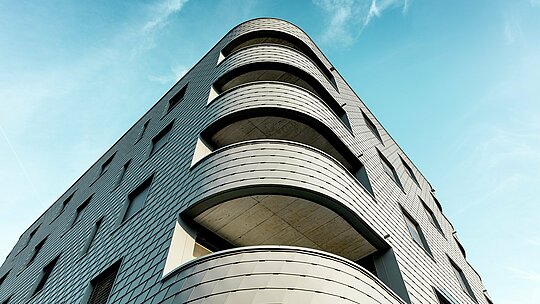 Vue dynamique du dessous d'un immeuble résidentiel et commercial moderne, caractérisé par un angle arrondi saisissant et des balcons aux lignes fluides. La façade, aux allures d'écailles, réalisée en bardage PREFA P.10 Gris souris, enveloppe le bâtiment tel une peau d'aluminium finement travaillée, soulignant la douceur de ses courbes. L'homogénéité de la surface met en valeur la pureté des lignes, tandis que la légèreté et la résistance aux intempéries de l'aluminium PREFA garantissent une stabilité dimensionnelle maximale, une grande durabilité et un entretien minimal, même pour les courbes les plus prononcées – un véritable chef-d'œuvre architectural se détachant sur un ciel d'un bleu éclatant.