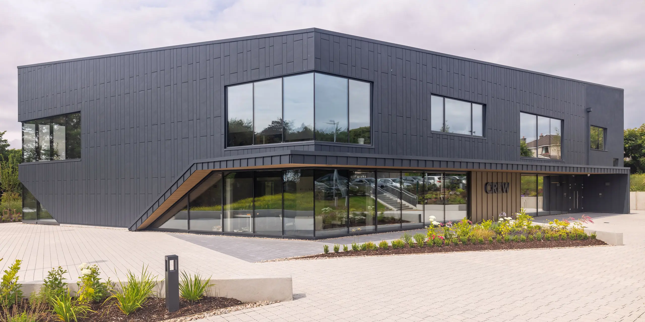 Vue frontale du bâtiment CREW à Galway, en Irlande, avec une façade moderne en aluminium PREFA en Siding.X et PREFALZ en anthracite P.10. Les lignes inclinées et les grandes fenêtres caractérisent la conception architecturale sophistiquée du bâtiment. Le bardage en aluminium anthracite combiné confère au bâtiment une esthétique élégante et contemporaine, complétée par un aménagement paysager bien entretenu.