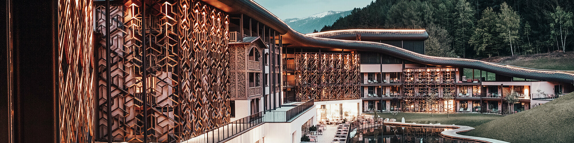 Prise de vue au crépuscule, une lumière chaude éclaire la façade en bois et le toit en aluminium. On aperçoit au rez-de-chaussée la terrasses et ses chaises-longues au bord du lac, en arrière-plan la forêt est vert sombre.