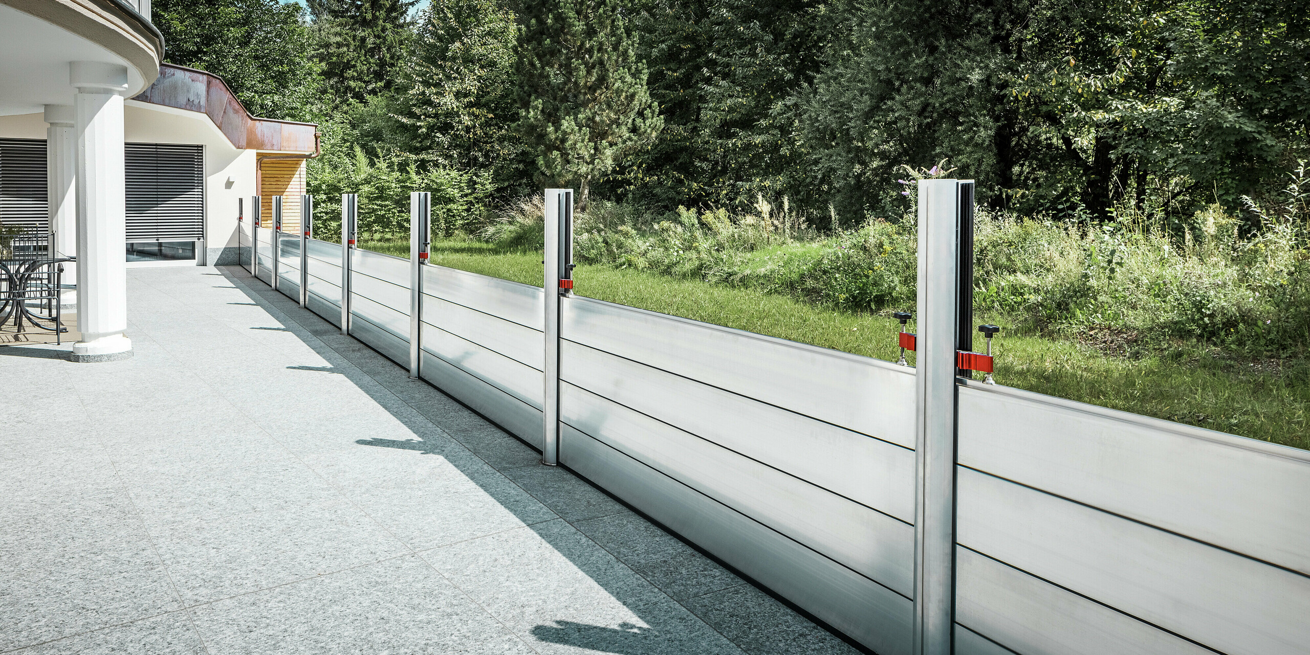 Vue latérale du mur de protection contre les crues PREFA entièrement installé le long de la terrasse d'un immeuble résidentiel au Tyrol. Les batardeaux robustes en aluminium sont fixées entre des supports verticaux dans des douilles de sol et protègent la propriété des inondations potentielles provenant de la zone naturelle adjacente. Le système impressionne par sa finition soignée, sa grande stabilité et son intégration discrète dans l'architecture moderne.