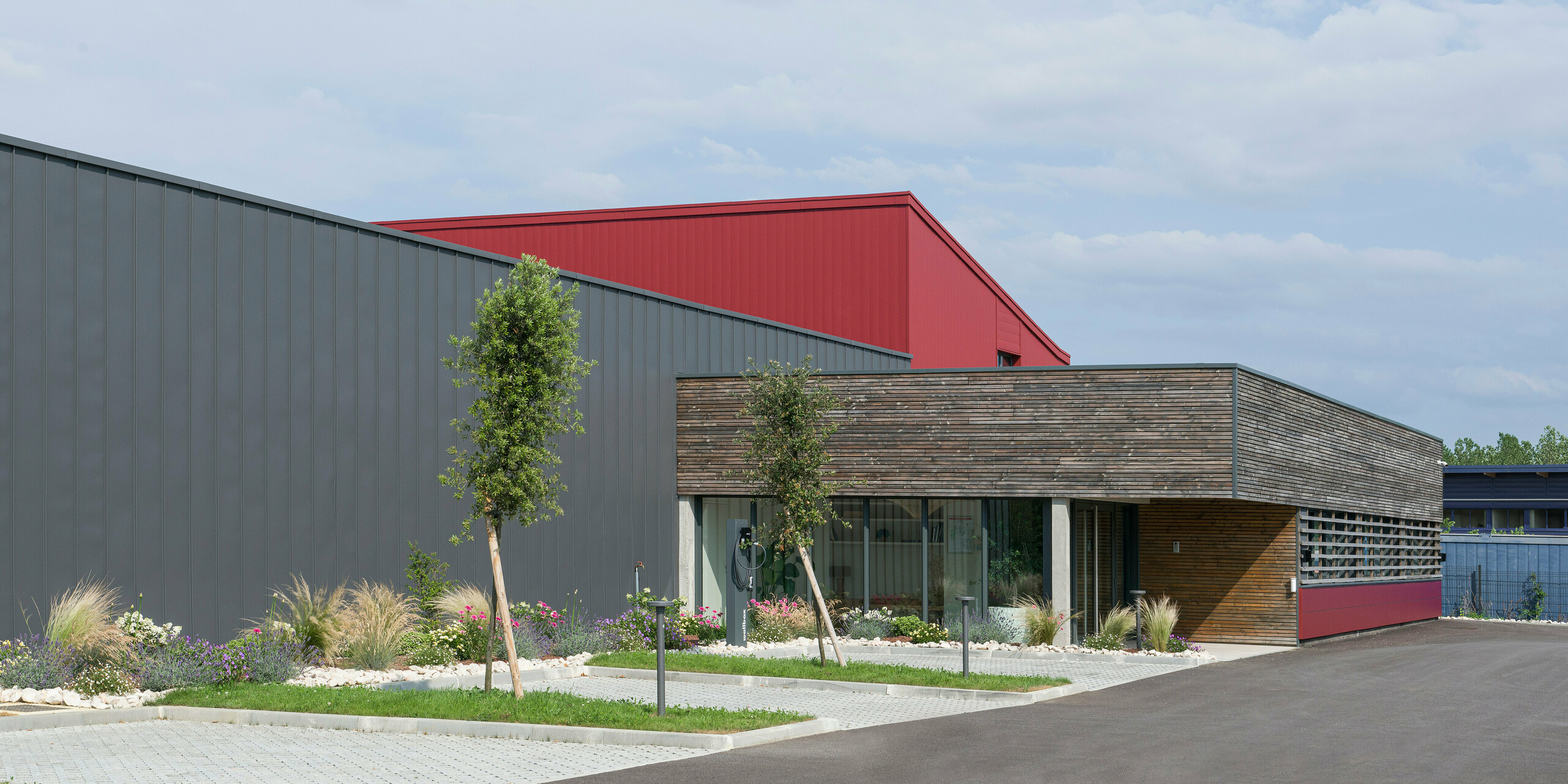 L’image montre un bâtiment moderne avec une combinaison frappante d’éléments de design industriel et naturel. La façade combine des éléments en aluminium gris foncé et rouge oxyde qui apportent dynamisme et structure. Les revêtements PREFA rouge oxyde apportent une touche colorée. La partie basse du bâtiment, dotée d'un mur revêtu de bois et de grandes fenêtres, crée une atmosphère chaleureuse et invitante, évoquant un espace à usage de bureau. Le jardin bien entretenu devant le bâtiment véhicule une image respectueuse de l'environnement et durable. La combinaison de ces éléments reflète le désir de combiner des méthodes de construction traditionnelles avec des matériaux et des conceptions modernes et durables.