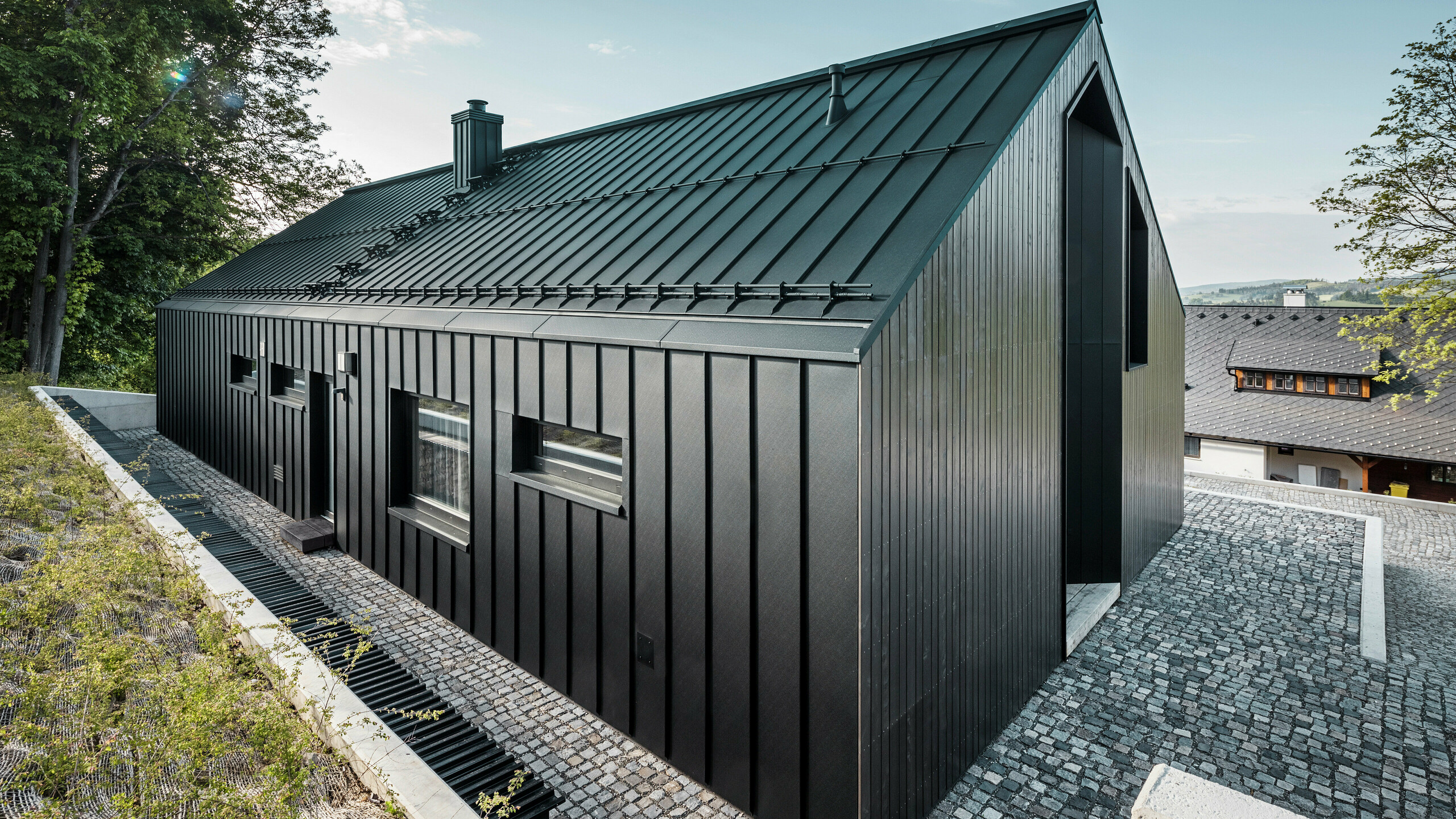 Blick von schräg vorne auf den Eingangsbereich bzw. die Gebäuderückseite eines modernen Einfamilienhauses in Kuncice (Tschechien). Die Front des Gebäudes ist mit einer dunklen Holzfassade verkleidet. Die Rückseite sowie das Dach des Hauses sind mit einem Stehfalzsystem von PREFA ummantelt. Das Dach- und Fassadensystem aus Aluminium ist in der edlen Farbe P.10 Schwarz gehalten. Am rechten Bildrand ist ein Nachbarhaus zu sehen. Links hinter dem Gebäude ist Wald.