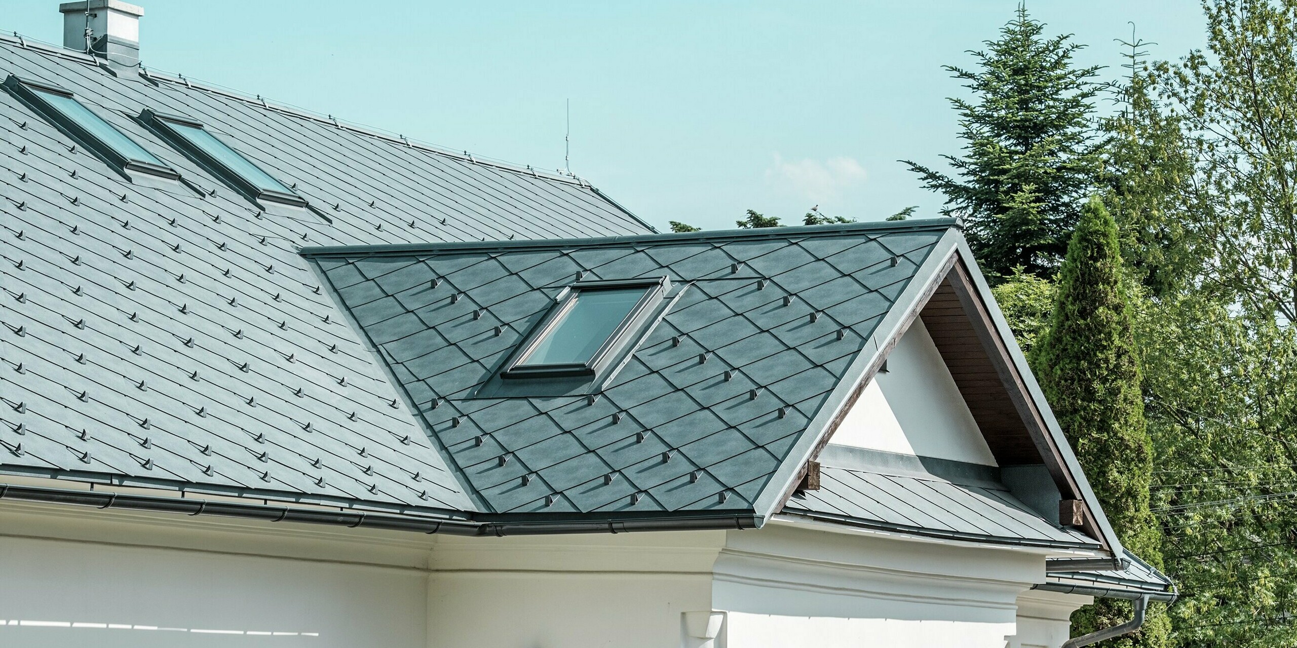 L'image présente une coupe détaillée d'une maison d'artisan à Klin, en Slovaquie. La toiture en losanges PREFA 44 x 44, coloris Gris Pierre P.10, offre un aspect moderne et attrayant, en parfaite harmonie avec l'architecture traditionnelle du bâtiment. Les fenêtres de toit, de dimensions généreuses, sont particulièrement remarquables : elles inondent de lumière l'étage et contribuent à la beauté du design. L'élégant toit à pignon, surmonté d'une lucarne au-dessus de l'entrée, impressionne par sa durabilité, sa légèreté et sa résistance, ce qui en fait un choix idéal pour une rénovation durable.