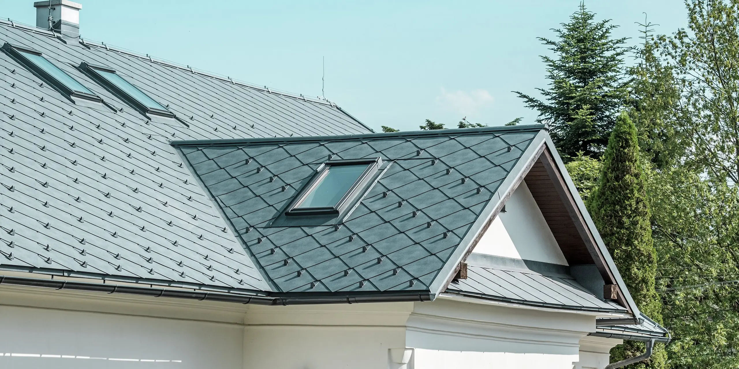 L'image présente une coupe détaillée d'une maison d'artisan à Klin, en Slovaquie. La toiture en losanges PREFA 44 x 44, coloris Gris Pierre P.10, offre un aspect moderne et attrayant, en parfaite harmonie avec l'architecture traditionnelle du bâtiment. Les fenêtres de toit, de dimensions généreuses, sont particulièrement remarquables : elles inondent de lumière l'étage et contribuent à la beauté du design. L'élégant toit à pignon, surmonté d'une lucarne au-dessus de l'entrée, impressionne par sa durabilité, sa légèreté et sa résistance, ce qui en fait un choix idéal pour une rénovation durable.