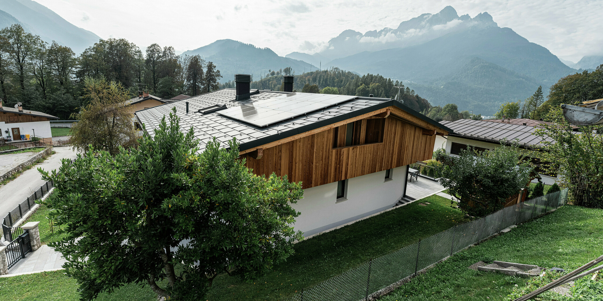 Arrière d'une maison familiale rénovée à Agordo, Italie. La nouvelle toiture a été recouverte de PREFA R.16 en P.10 anthracite. Le toit en aluminium durable offre une protection optimale contre les conditions météorologiques extrêmes et est équipé d'un système photovoltaïque durable. Le système de gouttière PREFA avec gouttière et tuyau de descente assure un drainage fiable de l'eau. La combinaison d'une façade en bois naturel, d'une façade en plâtre blanc et d'un toit en aluminium durable s'intègre harmonieusement dans le paysage alpin.