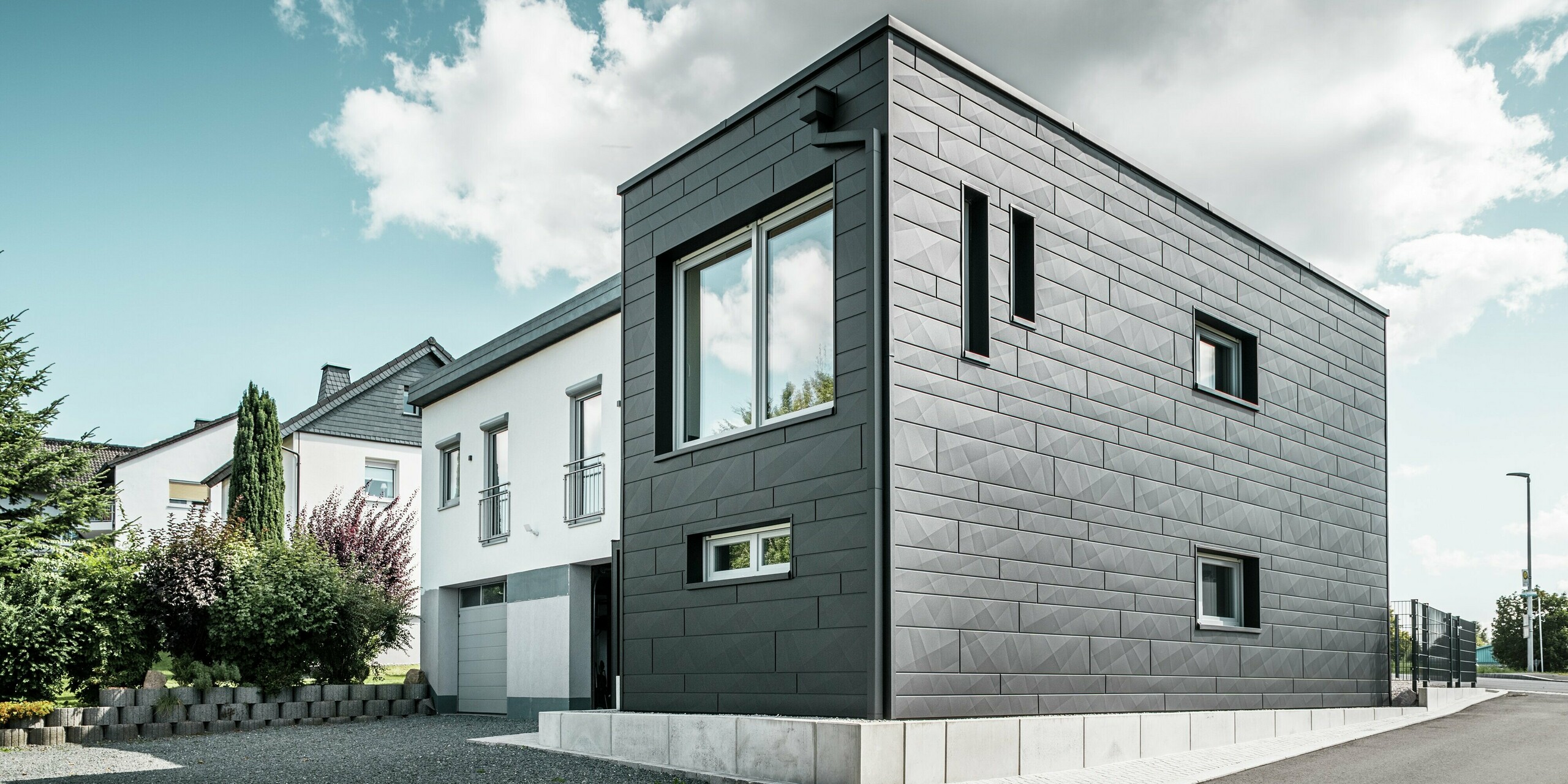 La façade élégante et moderne de la maison individuelle rénovée de Teuschnitz est ornée de panneaux Siding.X PREFA à la structure claire, coloris P.10 noir. La disposition géométrique des panneaux en aluminium de haute qualité confère au bâtiment une esthétique saisissante et intemporelle. Les fenêtres sont encadrées de panneaux composites en aluminium noir, complétant harmonieusement le design. Le système de gouttière PREFA intégré, avec son tuyau carré, offre une solution fonctionnelle et esthétique.
