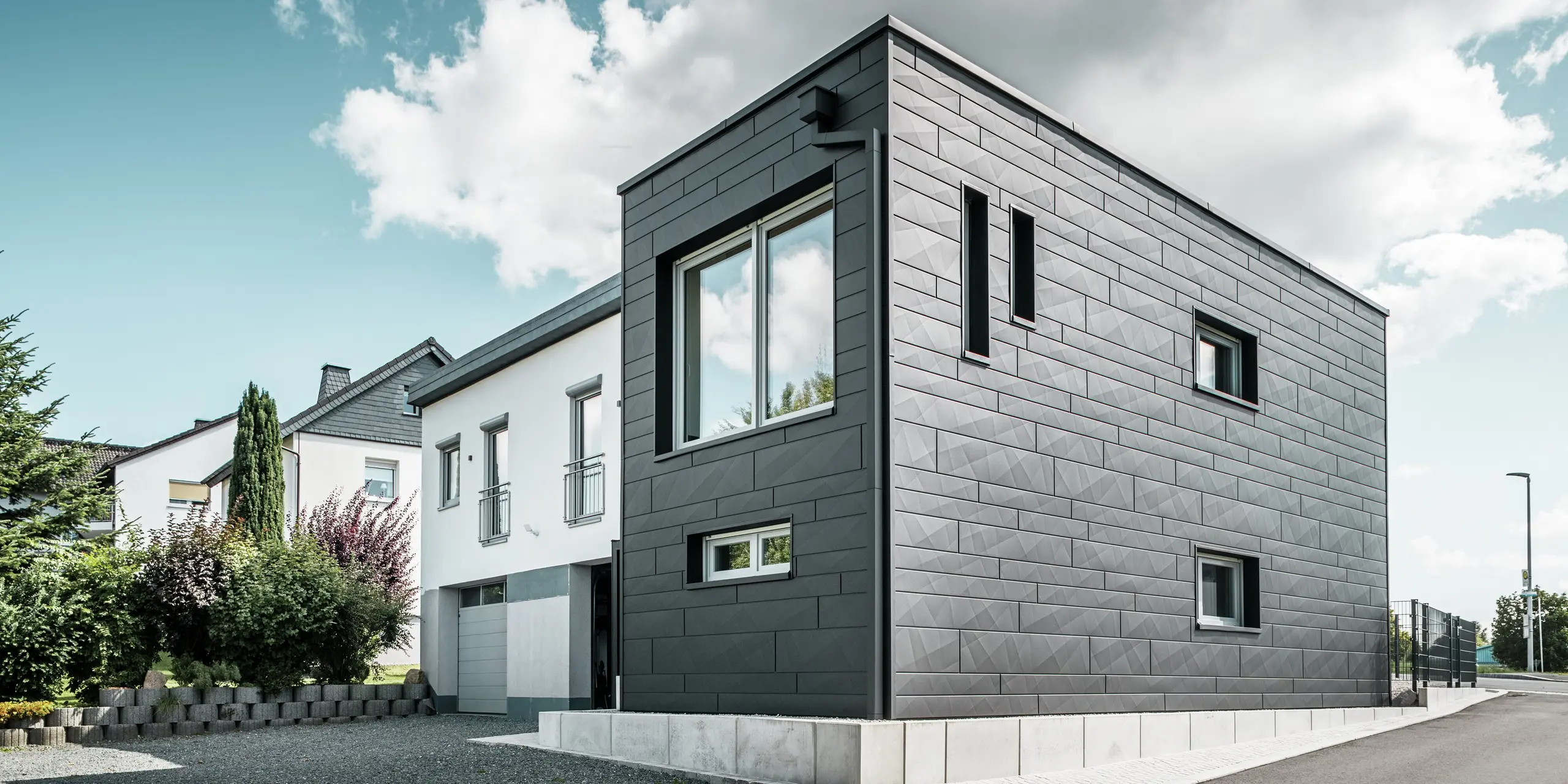 La façade élégante et moderne de la maison individuelle rénovée de Teuschnitz est ornée de panneaux Siding.X PREFA à la structure claire, coloris P.10 noir. La disposition géométrique des panneaux en aluminium de haute qualité confère au bâtiment une esthétique saisissante et intemporelle. Les fenêtres sont encadrées de panneaux composites en aluminium noir, complétant harmonieusement le design. Le système de gouttière PREFA intégré, avec son tuyau carré, offre une solution fonctionnelle et esthétique.