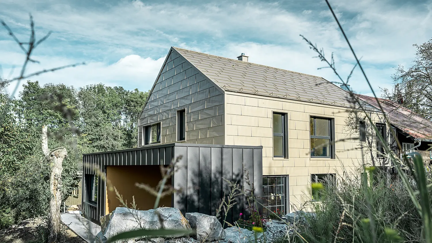 La toiture et la façade du bâtiment résidentiel sans avant-toit sont revêtues du panneau en aluminium FX.12 de PREFA couleur bronze. Juste à côté se trouve un garage à toit plat avec façade à joint debout.