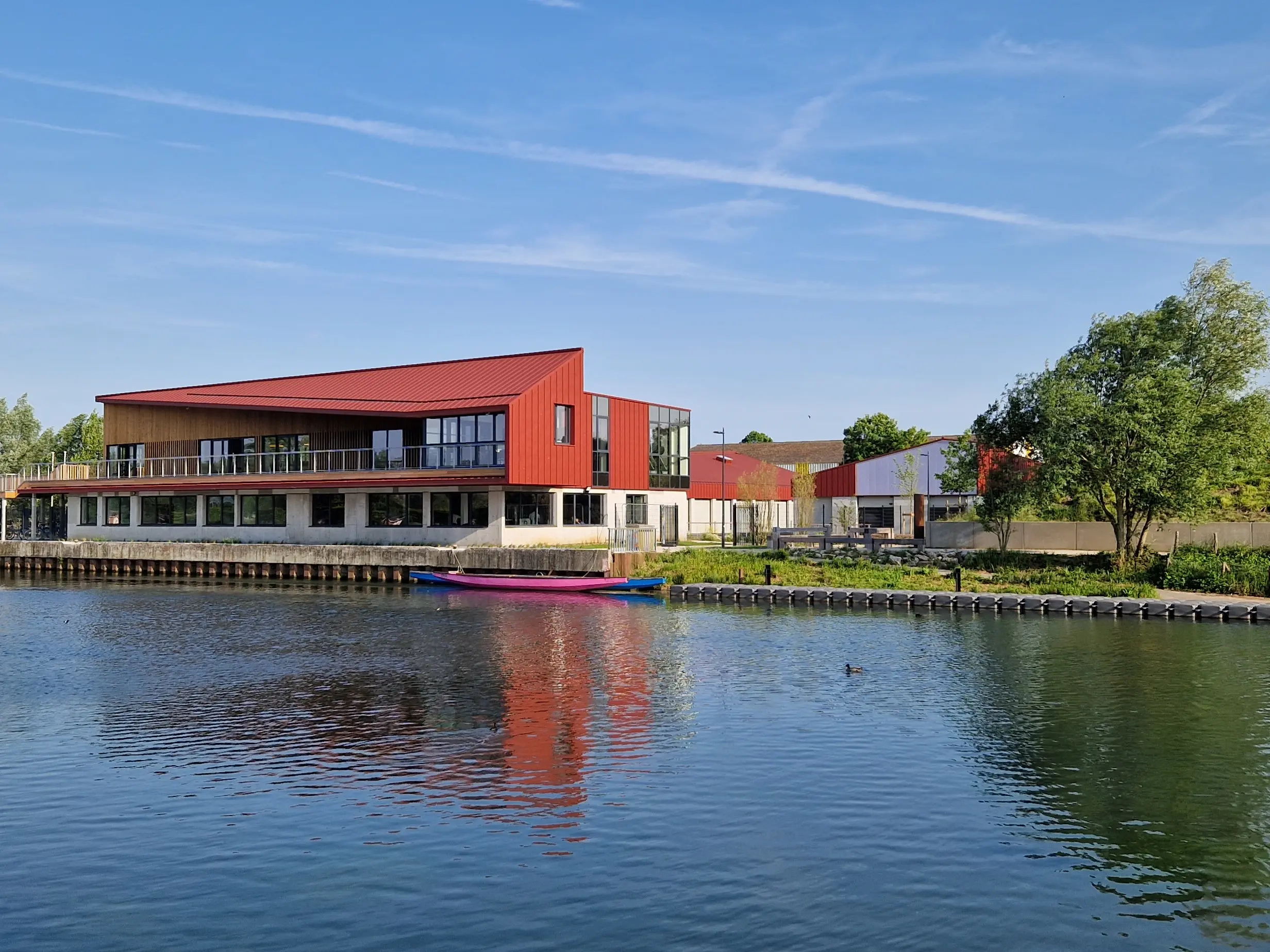 Vue éloignée du stade nautique d'Arras, la teinte rouge oxyde de l'aluminium Prefa se reflète dans la rivière