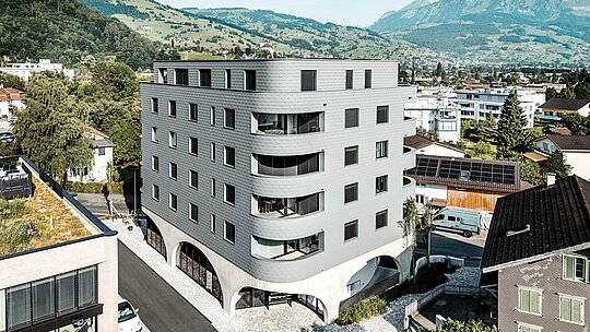 Vue aérienne de la Bernerhaus à Buchs, avec sa forme triangulaire saisissante et ses balcons aux courbes douces, nichée au cœur du paysage alpin. La façade continue en bardage PREFA gris souris P.10 enveloppe le bâtiment comme une peau d'aluminium aux allures d'écailles, créant un jeu de lumière et de textures saisissant. Au-dessus du socle en béton massif orné d'arcades, la façade légère et résistante aux intempéries en aluminium PREFA se distingue par sa précision, sa durabilité et son faible besoin d'entretien – un élément architectural moderne et remarquable dans le paysage urbain.