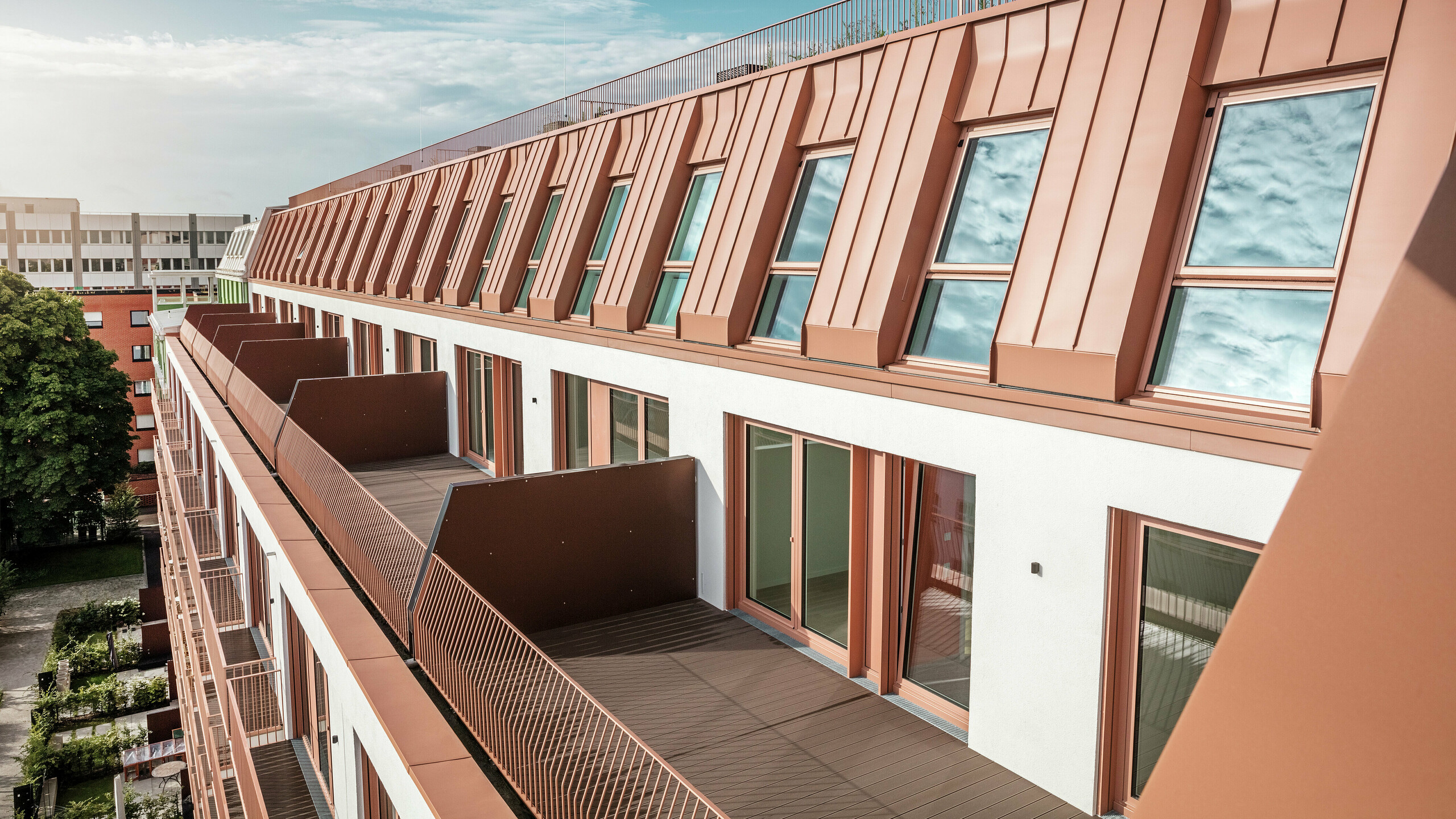 Vue détaillée de la conception moderne du toit et de la façade du projet résidentiel Van B à Munich avec le toit en aluminium PREFALZ de haute qualité en IGP Orange HDP de PREFA. Le traitement précis du matériau durable et nécessitant peu d'entretien offre une protection optimale contre les éléments et confère au bâtiment un aspect uniforme et élégant. Les généreuses fenêtres et balcons offrent beaucoup de lumière naturelle et créent un environnement de vie ouvert et habitable avec une sophistication architecturale.