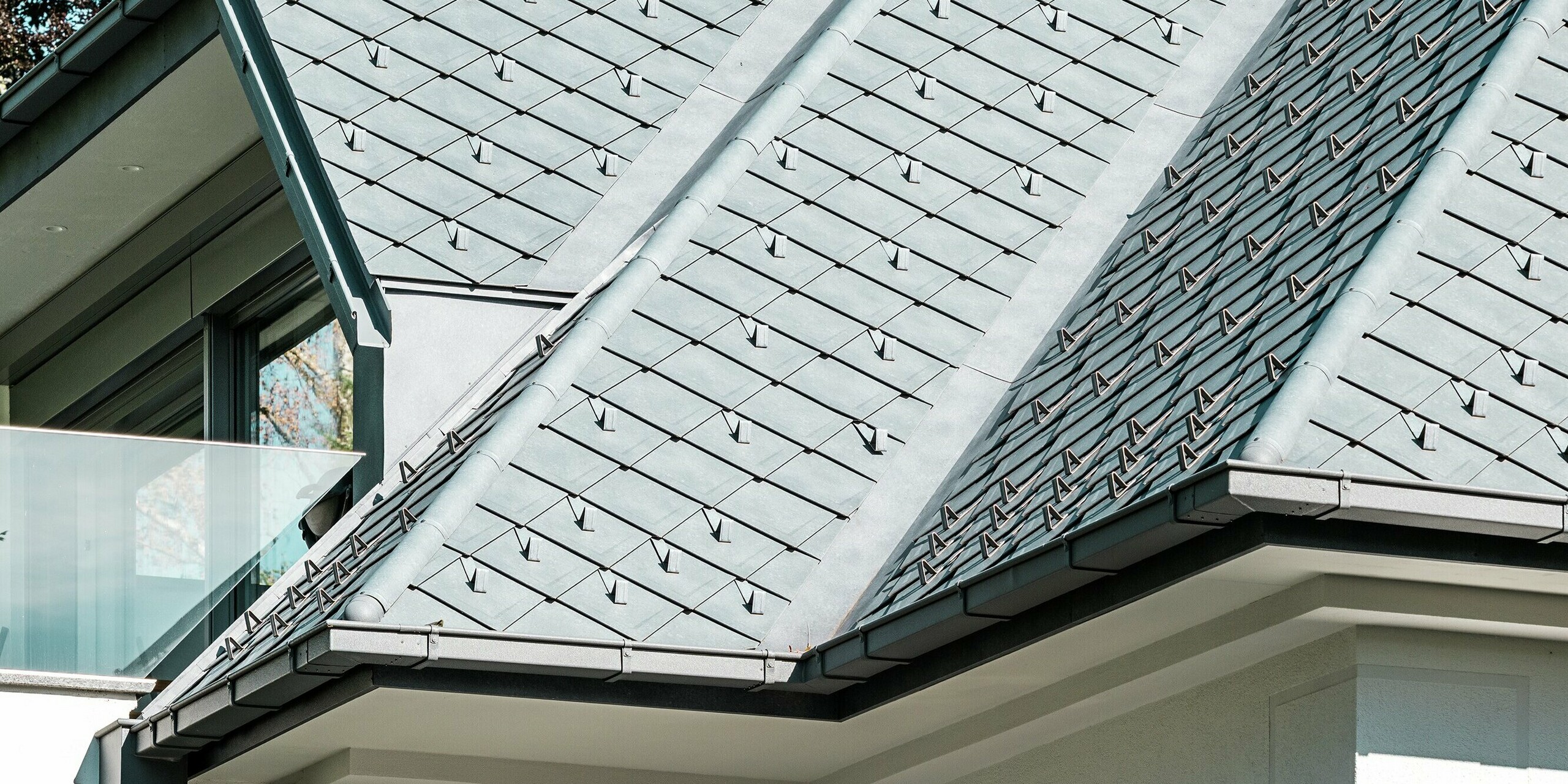 Gros plan sur un immeuble d'appartements moderne doté d'une élégante toiture en losanges PREFA 29 x 29 cm, coloris Gris Pierre P.10. Des pare-neige intégrés assurent une sécurité accrue en hiver. L'élégant système d'évacuation des eaux pluviales PREFA, avec sa gouttière et sa descente, complète l'ensemble et garantit une évacuation efficace. L'alliance de matériaux de haute qualité et d'une conception soignée confère à l'immeuble une esthétique soignée, tout en le rendant fonctionnel et durable.