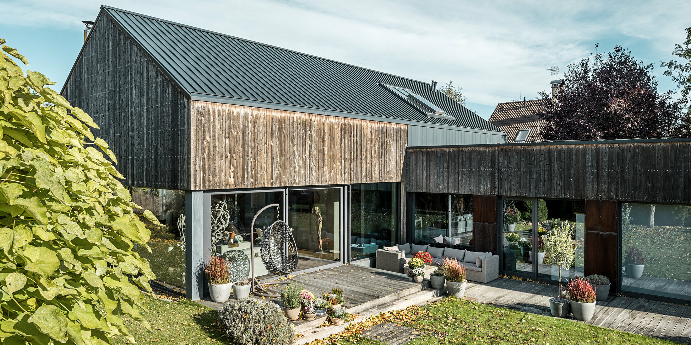 Maison individuelle avec toit à pignon, recouvert avec les joints debout double agrafe PREFALZ en anthracite et façade en bois patiné. Avec belle terrasse en bois et grandes fenêtres au rez-de-chaussée.