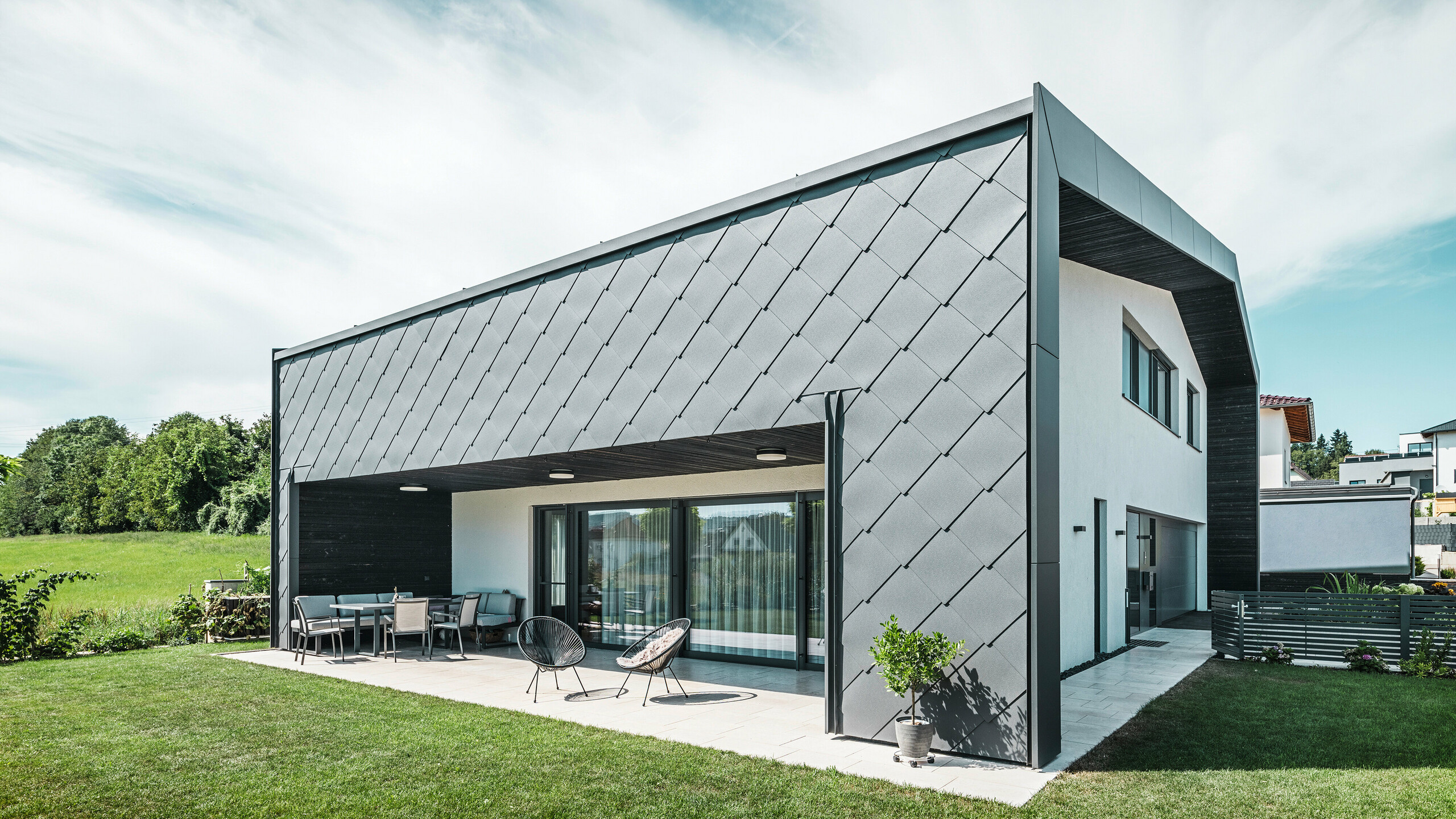 Jardin bien entretenu d'une maison unifamiliale à Aschach. Le bâtiment est élégamment recouvert de losanges de façade 44x44 gris sombre. La terrasse couverte offre un coin salon confortable avec vue sur la pelouse verte luxuriante. Le revêtement élégant en aluminium associé à des surfaces en plâtre blanc garantit une architecture moderne et résistante aux intempéries. De grandes fenêtres laissent entrer beaucoup de lumière du jour à l'intérieur et créent une atmosphère ouverte et lumineuse.