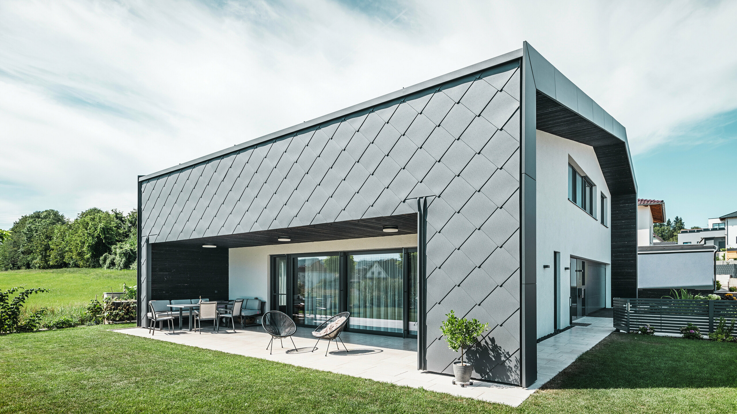 Jardin bien entretenu d'une maison unifamiliale à Aschach. Le bâtiment est élégamment recouvert de losanges de façade 44x44 gris sombre. La terrasse couverte offre un coin salon confortable avec vue sur la pelouse verte luxuriante. Le revêtement élégant en aluminium associé à des surfaces en plâtre blanc garantit une architecture moderne et résistante aux intempéries. De grandes fenêtres laissent entrer beaucoup de lumière du jour à l'intérieur et créent une atmosphère ouverte et lumineuse.