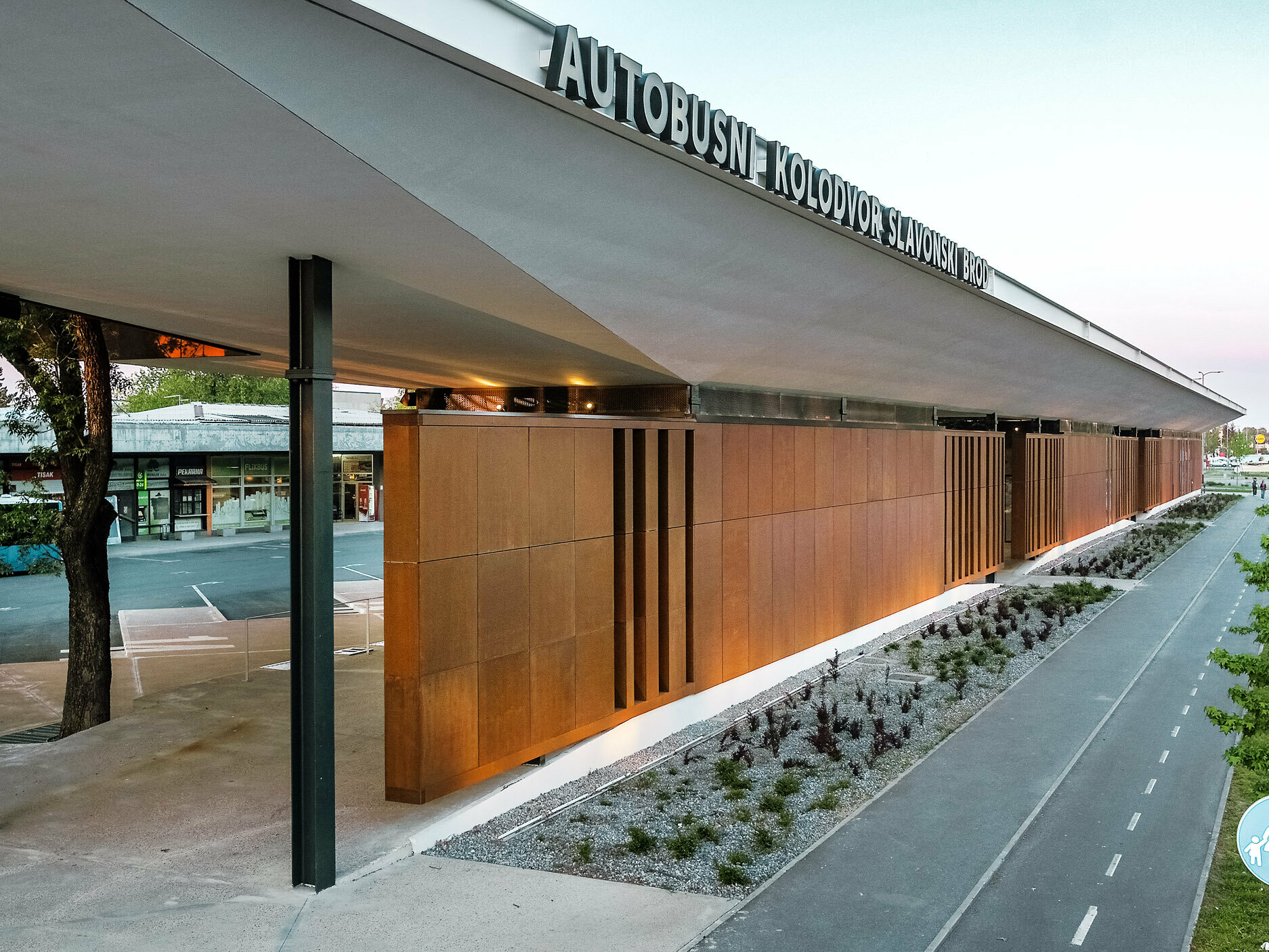 La photo montre le hall moderne de la gare routière « Autobusni Kolodvor Slavonski Brod » en Croatie. Le hall de gare présente une façade remarquable de panneaux bruns soutenus par des colonnes noires. Le toit Prefalz blanc de PREFA s'étire sur toute la structure, protégeant les espaces situés en dessous. Au premier plan, on peut voir une piste cyclable flanquée d'un espace vert bien entretenu et de jeunes arbres. À l'arrière-plan, on distingue un bus bleu clair, ainsi que divers magasins.