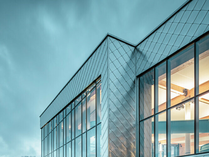 Dans le cliché, on peut apercevoir les losanges de façade 44x44 en aluminium naturel qui recouvre la façade du centre Hardangerbadet à Oystese. Des nuages menaçant se trouvent en arrière plan. 