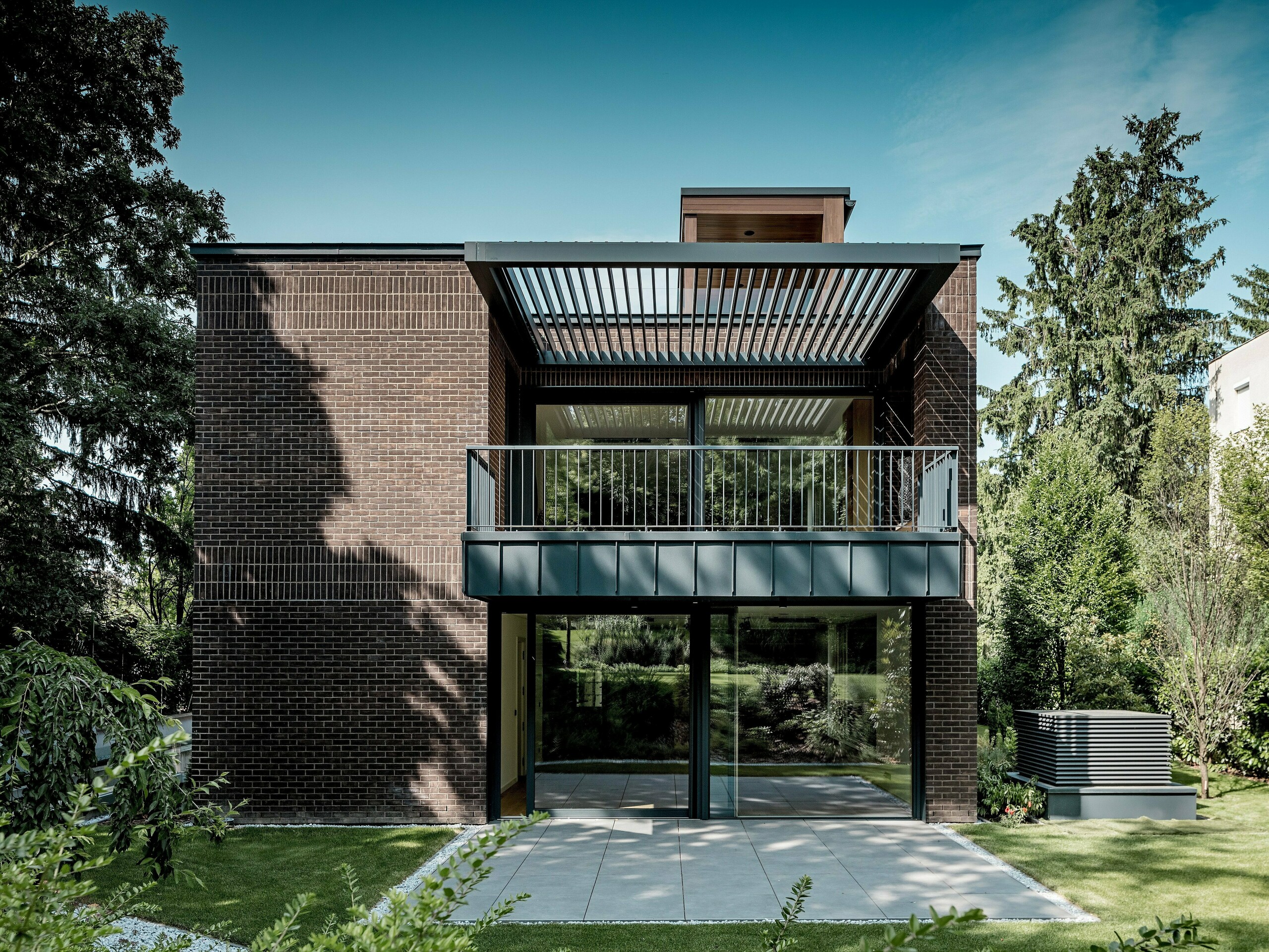 La photographie révèle la façade côté jardin d'une villa moderne et multigénérationnelle à Budapest. Sa structure épurée associe briques rouges foncées et aluminium PREFALZ P.10 anthracite. Le vaste balcon, avec sa délicate charpente métallique et ses persiennes, crée un jeu d'ombres et de lumières captivant. De grandes baies vitrées ouvrent l'intérieur sur le jardin luxuriant. La précision d'exécution des détails en aluminium souligne la durabilité, la résistance aux intempéries et la clarté architecturale des solutions PREFALZ, conférant au bâtiment un caractère saisissant et intemporel.