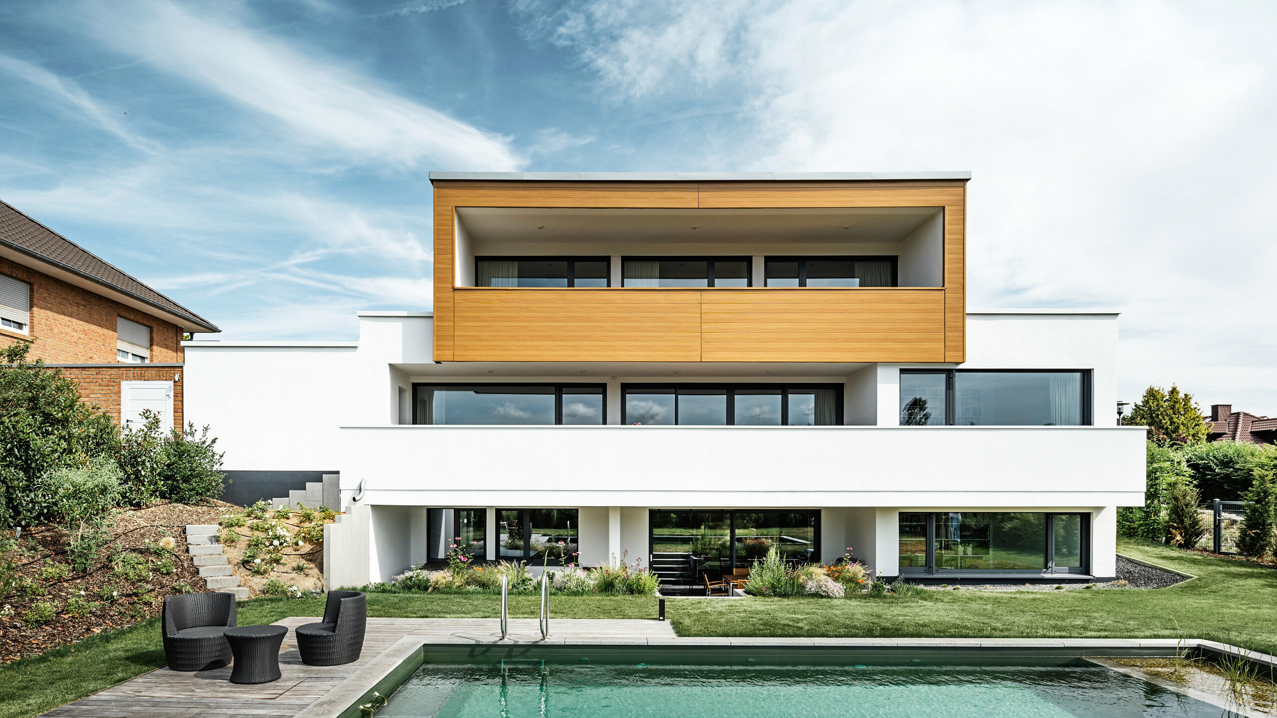 Die Rückseite eines Einfamilienhauses in Usingen, Deutschland. Am unteren Bildrand ist der gepflegte Pool und Garten inkl. Outdoor-Sitzmöbel zu sehen. Das oberste Geschoss des Gebäudes wurde mit PREFA Sidings in Eiche natur umhüllt. Der Gebäudesockel wurde mit einer weißen Putzfassade ausgestattet.