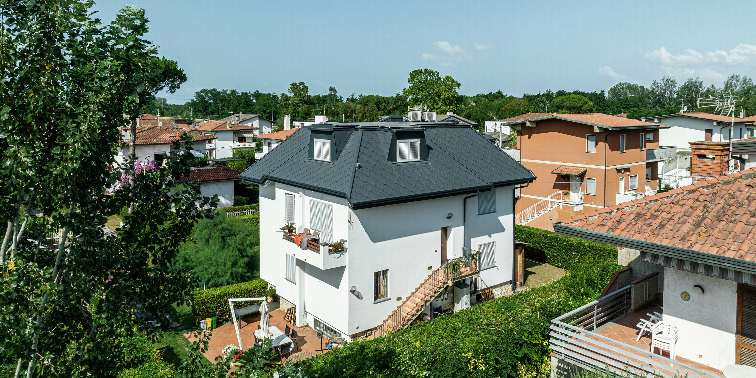 Maison individuelle élégante à Montignoso vue d'une perspective surélevée. Le toit en losanges PREFA 44 × 44 en P.10 gris pierre confère au toit une apparence élégante et impressionne par sa durabilité et sa résistance extrême aux intempéries. La toiture sombre forme un beau contraste avec la façade blanche. Les lucarnes s'intègrent harmonieusement à la structure du toit.