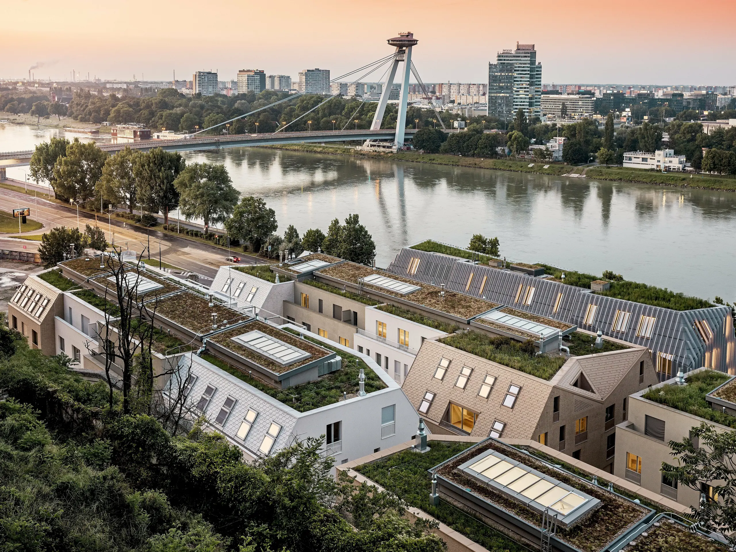 Une vue spectaculaire sur le quartier de Vydrica à Bratislava, avec les rives du Danube et son pont remarquable baignés par la lumière du soir : la toiture aux multiples facettes, composée de toits végétalisés, de puits de lumière parfaitement intégrés et de toits à forte pente revêtus de bardeaux d’aluminium PREFA blancs, brun sable et bronze, offre un spectacle visuel saisissant. La pureté des lignes et l’harmonie des couleurs unissent avec brio l’architecture à son environnement. L’aluminium PREFA se distingue par son extrême durabilité, sa légèreté, sa grande résistance aux intempéries et sa conception flexible – idéal pour des solutions de toiture complexes et durables.