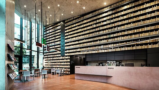 L'image révèle l'intérieur moderne et élégant de la bibliothèque d'Olomouc, dominé par son impressionnante bibliothèque murale. Les étagères du sol au plafond créent un effet saisissant, tandis que le comptoir d'accueil, à la fois simple et raffiné, réalisé en béton et bois de haute qualité, instaure une atmosphère chaleureuse. La pureté des lignes et la précision de l'éclairage, qui s'intègrent harmonieusement à l'ensemble, sont particulièrement remarquables. L'utilisation de panneaux composites en aluminium PREFABOND et d'autres matériaux haut de gamme pour la façade extérieure garantit une structure durable et facile d'entretien. Cette architecture allie avec brio fonctionnalité et esthétique.