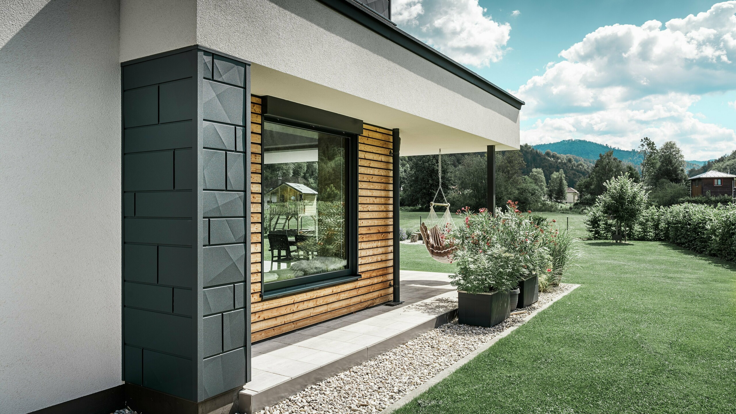 Vue partielle d'une maison individuelle récemment construite. Différents matériaux ont été utilisés pour la façade. La façade en crépi blanc est mise en valeur par des éléments en aluminium PREFA Siding.X anthracite ainsi que par des panneaux de bois horizontaux qui entourent une grande fenêtre. Une chaise suspendue est accrochée à la terrasse.