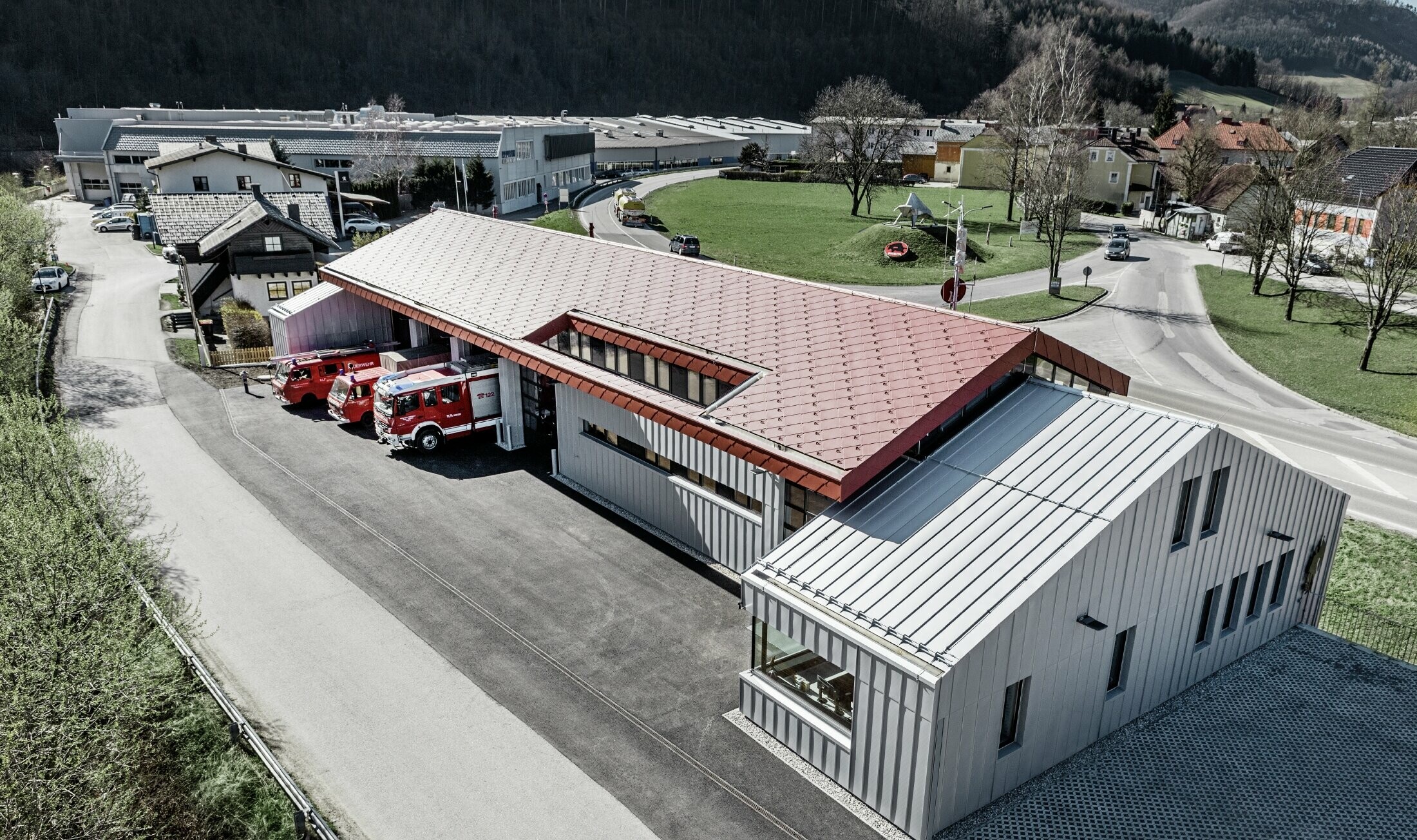 Vue aérienne de la caserne de sapeurs pompiers de l’entreprise à Marktl/Lilienfeld. La toiture et la façade de la partie gauche du bâtiment ont été entièrement habillées avec la tuile 44 couleur rouge oxyde. La partie droite est recouverte d’une enveloppe en aluminium réalisée avec Prefalz couleur argent métallisé.