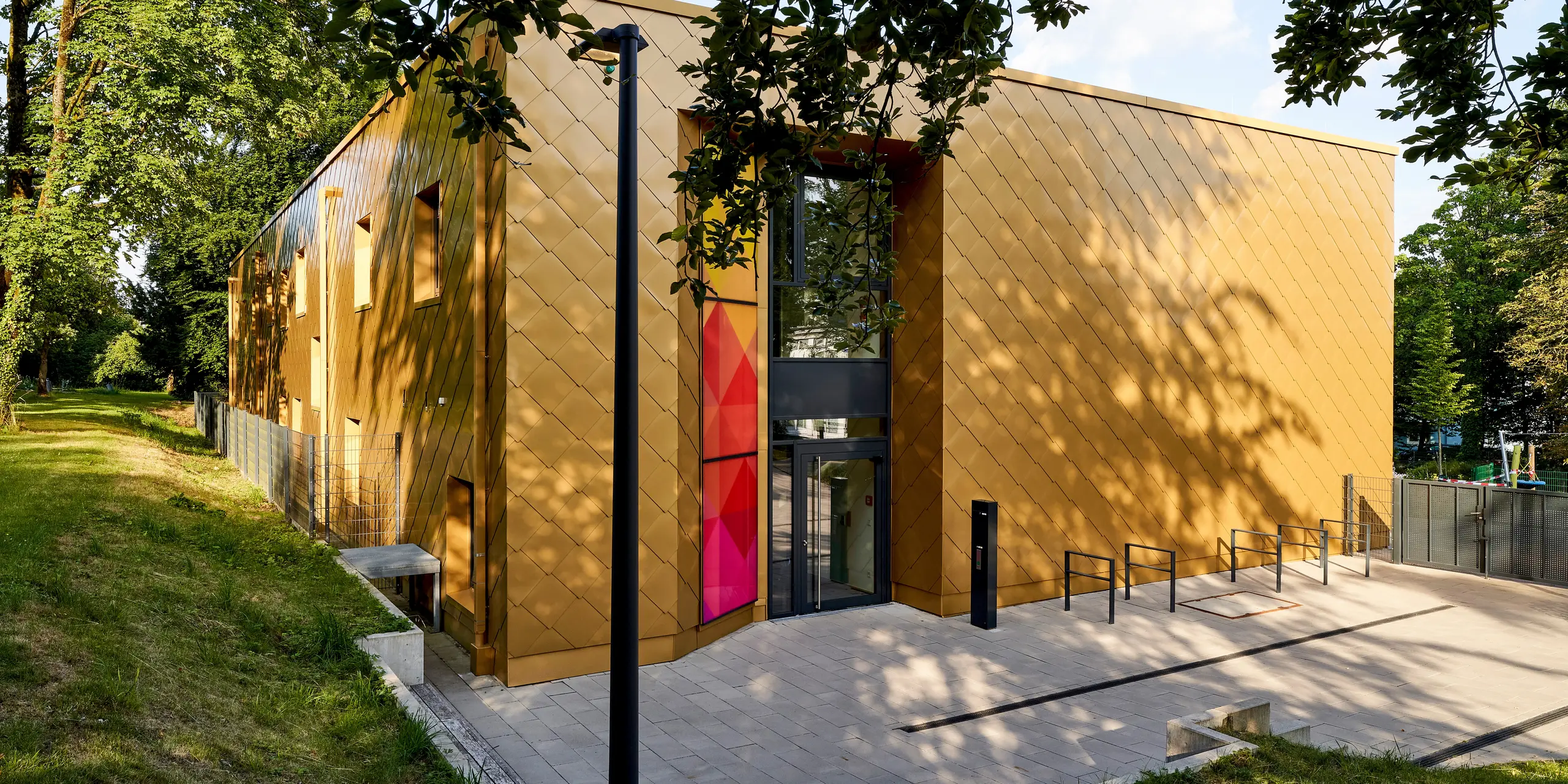 Une architecture exceptionnelle, avec une façade en aluminium PREFA couleur or et une structure murale précise en forme de losange, caractérise ce paysage impressionnant. Sous la canopée de feuillage, de vibrants jeux d'ombre et de lumière soulignent la silhouette saisissante du bâtiment, tandis qu'une entrée aux couleurs chatoyantes attire immédiatement le regard. Les losanges d'aluminium disposés en diagonale confèrent à la façade profondeur et dynamisme. Les produits en aluminium PREFA impressionnent ici par leur durabilité, leur résistance aux intempéries et l'éclat de leurs couleurs – une solution durable et sans entretien pour une architecture moderne en harmonie avec la nature.