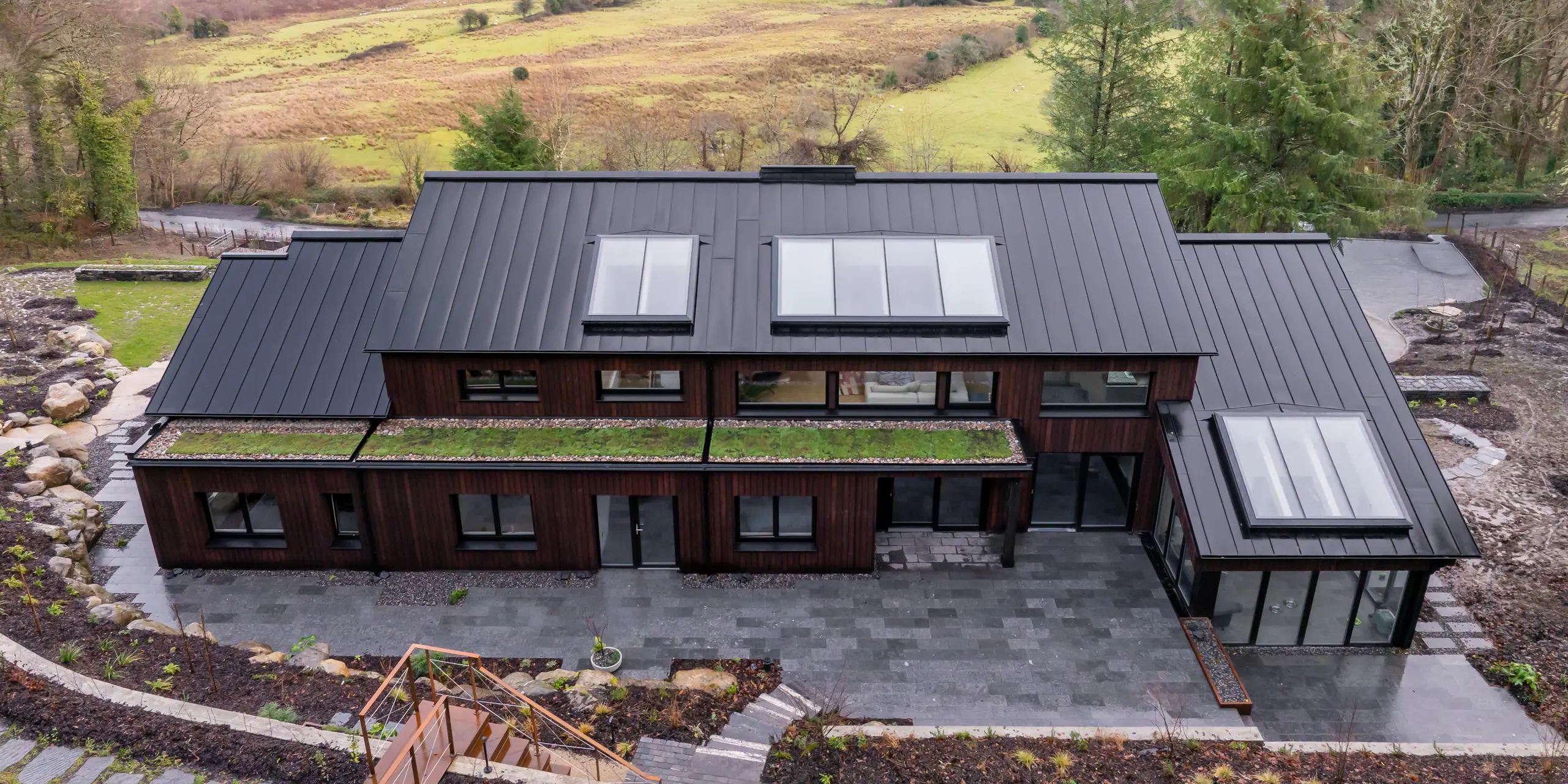 À Sligo, en Irlande, une maison individuelle moderne se distingue par sa façade en bois et son élégante toiture en aluminium PREFALZ P.10 Noir. La structure à joints debout épurée met en valeur l'architecture et s'intègre harmonieusement au vaste paysage de rivières et de collines. De larges baies vitrées et des puits de lumière apportent une touche d'originalité. Durable et résistante à la corrosion, la toiture PREFA impressionne par sa robustesse face aux intempéries et offre une solution élégante et facile d'entretien.