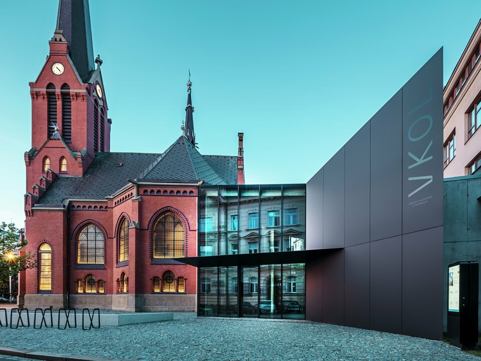 L'image révèle l'interaction remarquable entre l'église rouge historique et le nouveau bâtiment moderne d'Olomouc. L'église, avec ses fenêtres gothiques caractéristiques et son clocher imposant, constitue la colonne vertébrale historique de l'ensemble, tandis que l'extension moderne, avec son élégante façade en panneaux composites en aluminium PREFABOND noirs, représente une réponse architecturale contemporaine. De larges baies vitrées offrent des vues sur l'intérieur du nouveau bâtiment, où les reflets dans les fenêtres créent un jeu subtil entre ancien et nouveau. La précision de la fabrication des panneaux PREFABOND souligne l'esthétique moderne, tandis que l'intégration harmonieuse dans le contexte historique impressionne autant par sa fonctionnalité que par son esthétique.