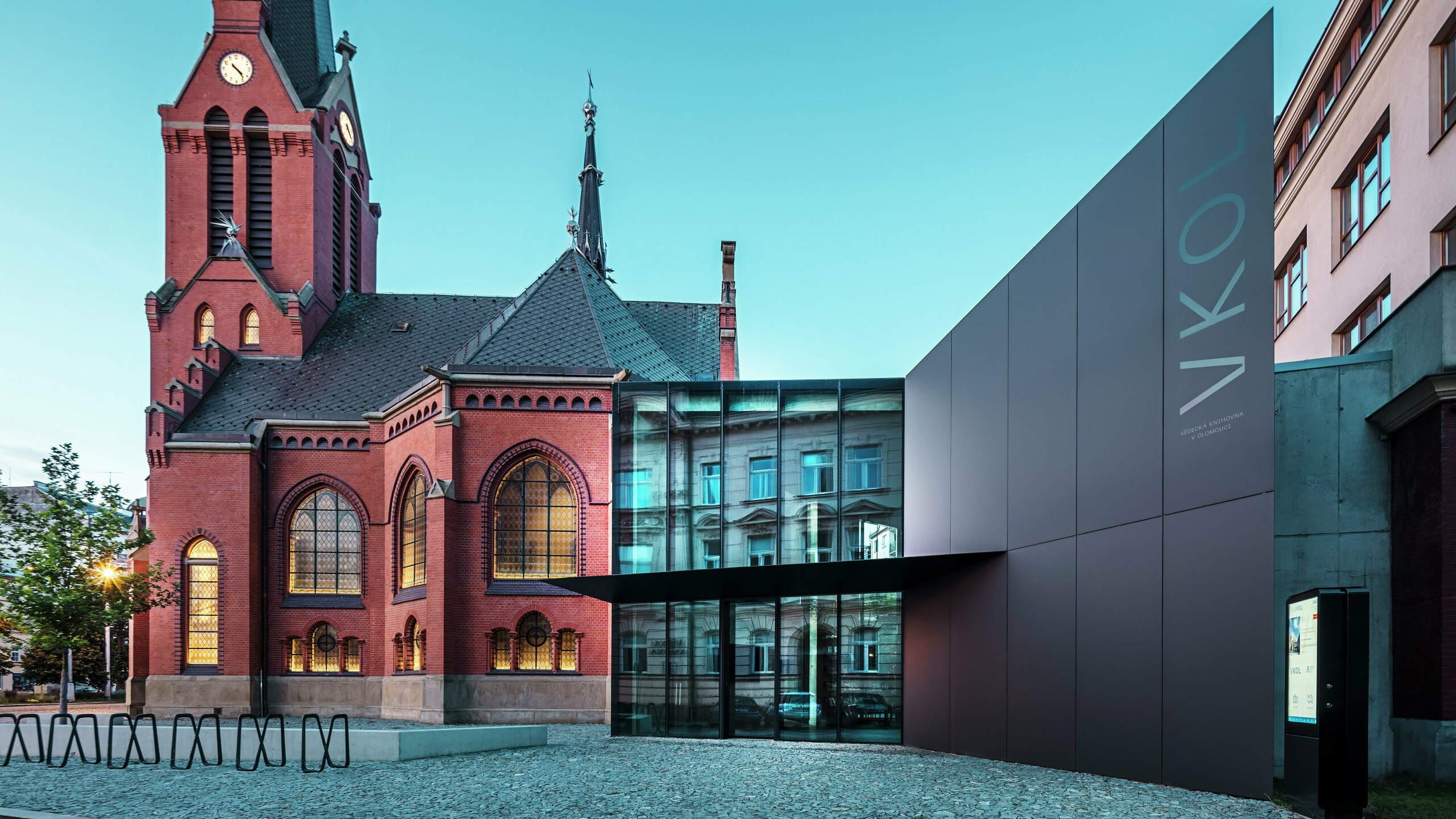 L'image révèle l'interaction remarquable entre l'église rouge historique et le nouveau bâtiment moderne d'Olomouc. L'église, avec ses fenêtres gothiques caractéristiques et son clocher imposant, constitue la colonne vertébrale historique de l'ensemble, tandis que l'extension moderne, avec son élégante façade en panneaux composites en aluminium PREFABOND noirs, représente une réponse architecturale contemporaine. De larges baies vitrées offrent des vues sur l'intérieur du nouveau bâtiment, où les reflets dans les fenêtres créent un jeu subtil entre ancien et nouveau. La précision de la fabrication des panneaux PREFABOND souligne l'esthétique moderne, tandis que l'intégration harmonieuse dans le contexte historique impressionne autant par sa fonctionnalité que par son esthétique.