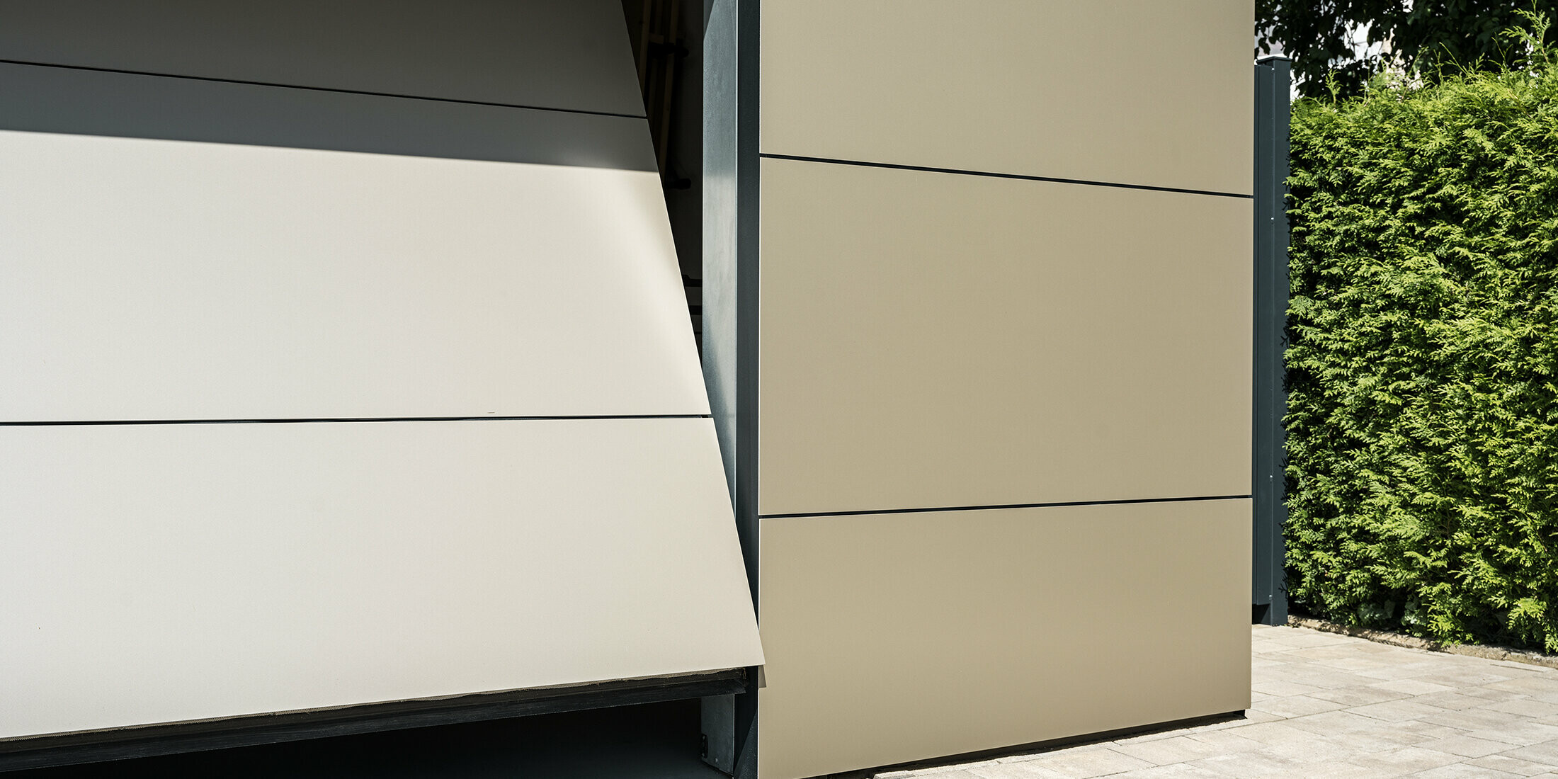 Nahaufnahme eines automatischen Garagentors. Die Garage und das automatische Garagentor sind mit bronzefarbenen Aluminium Verbundplatten verkleidet. Die präzise Verarbeitung spiegelt die Qualität der langlebigen Verbundplatten wider. Der schmale Öffnungsspalt gewährt einen kleinen Einblick in das Innere der Garage.