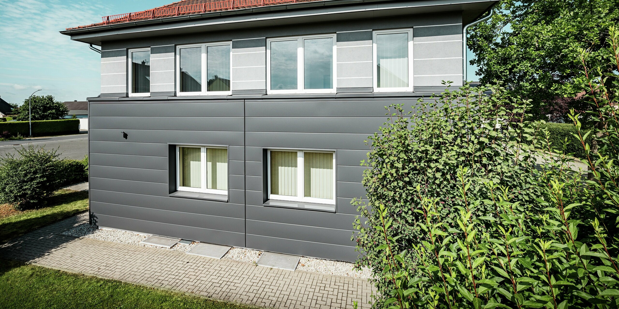 L'image présente la façade modernisée de la guilde des ramoneurs de Bindlach, revêtue de Sidings PREFA couleur Gris sombre P.10. Les lignes épurées des panneaux d'aluminium confèrent à la façade une esthétique contemporaine et attrayante, tandis que sa robustesse assure une excellente protection contre les intempéries. L'harmonieuse combinaison du gris foncé et de la végétation luxuriante donne au bâtiment un aspect frais et dynamique. Grâce à la durabilité et à la facilité d'entretien des matériaux, la façade conservera son aspect impeccable pendant de nombreuses années, garantissant ainsi un attrait esthétique durable.