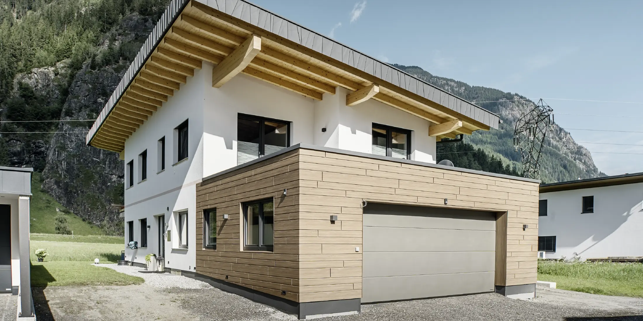 Maison individuelle moderne avec rénovation de façade : façade extérieure rénovée avec un revêtement horizontal aspect bois, combiné à des surfaces crépies en blanc, un revêtement de garage intégré et une architecture contemporaine dans un environnement alpin.