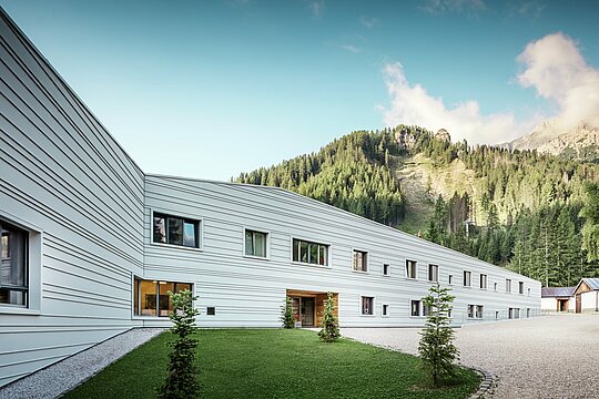 L'aluminium rencontre la précision : un dialogue harmonieux entre technique et nature. Dans le cadre impressionnant des Dolomites, le « Villaggio Alpino Tempesti » se présente avec des lignes claires et un design moderne. La façade en PREFALZ de couleur P.10 blanc Prefa est non seulement extrêmement résistante, mais sa couleur claire garantit également un microclimat agréable, car elle réfléchit les rayons du soleil. Les espaces verts et les éléments en bois à l'entrée du bâtiment militaire créent une atmosphère étonnamment accueillante.
