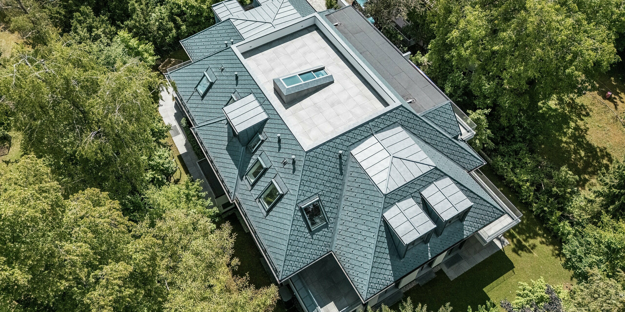 Cette photographie aérienne révèle un immeuble d'appartements moderne doté d'une remarquable toiture mansardée, recouverte de losanges PREFA de 29 x 29, coloris Gris Pierre P.10. Des lucarnes plates PREFALZ complètent la toiture, créant une silhouette structurée et distinctive qui s'intègre harmonieusement à la végétation environnante. La toiture plate centrale, accessible, renforce le caractère moderne de l'architecture. L'utilisation de produits en aluminium, durables et résistants aux intempéries, garantit des avantages à la fois esthétiques et fonctionnels.