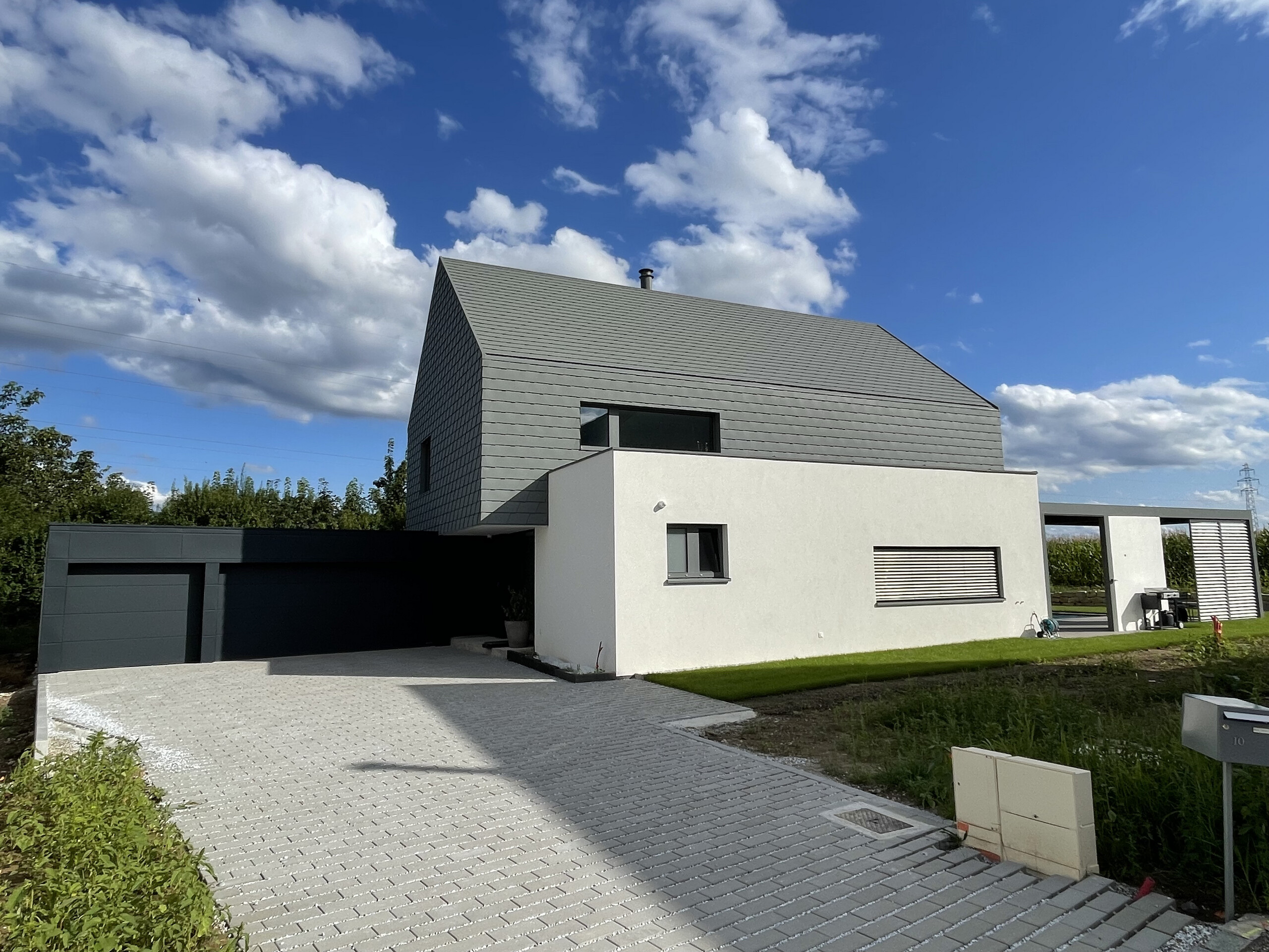 Vue éloignée de l'avant de cette maison moderne alsacienne. On aperçoit la cours pavée, le garage et les éléments en aluminium PREFA.