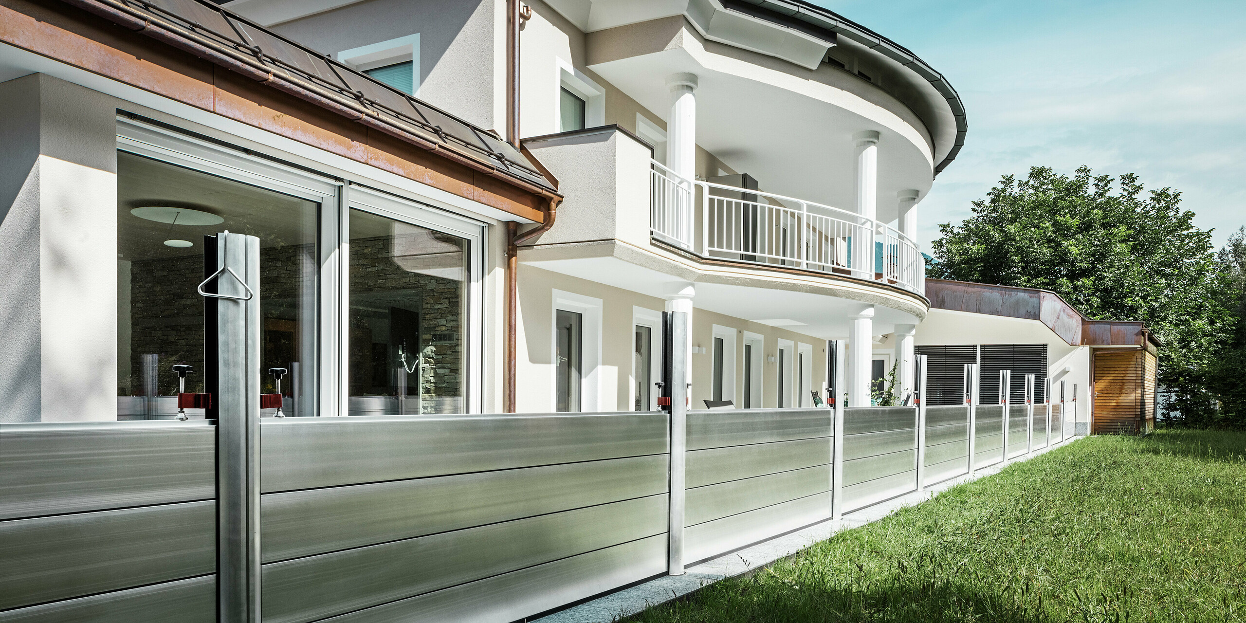 Vue d'un immeuble résidentiel moderne au Tyrol, doté d'un système de protection contre les crues PREFA installé le long de la terrasse. Les batardeaux robustes  en aluminium sont montées entre des profilés porteurs stables et protègent le bâtiment des inondations provenant des espaces verts et des cours d'eau adjacents. L'élégante maison, avec son balcon arrondi, ses colonnes blanches et ses grandes fenêtres, démontre comment une protection contre les inondations fonctionnelle peut s'intégrer harmonieusement à une architecture de qualité.
